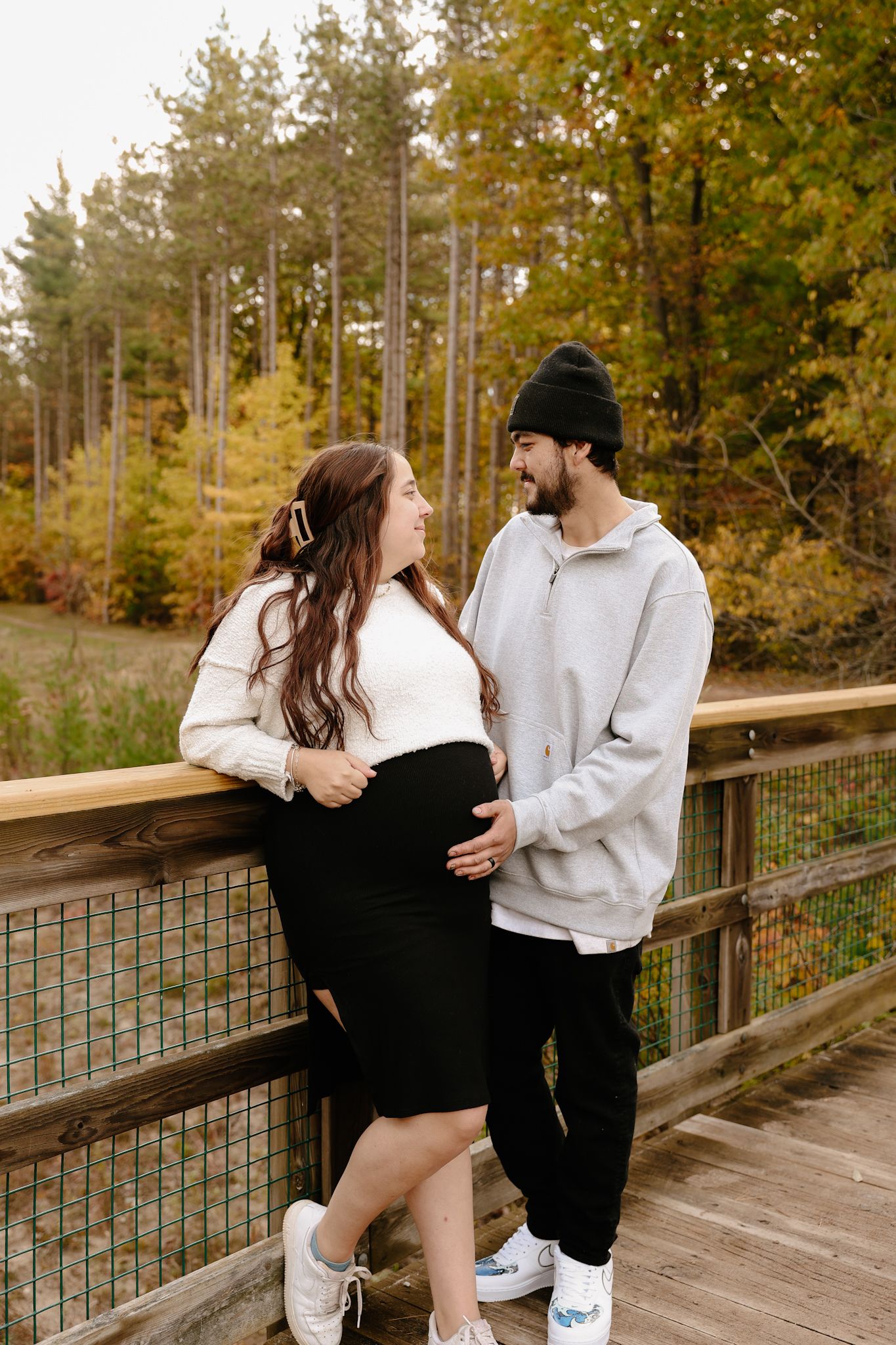 Pregnant couple sharing a tender moment on a wooden bridge surrounded by colorful autumn foliage.