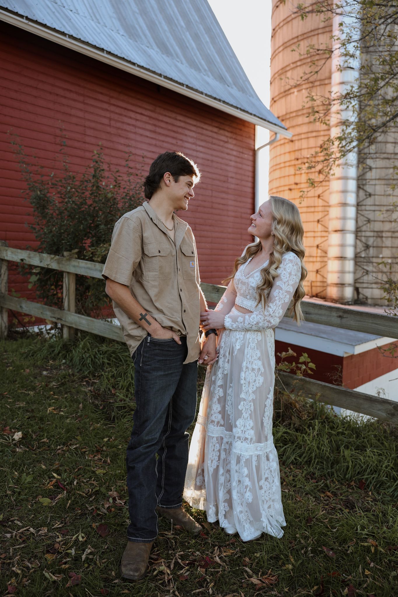 Young couple holding hands and smiling at each other beside a red barn and grain silos at sunset.