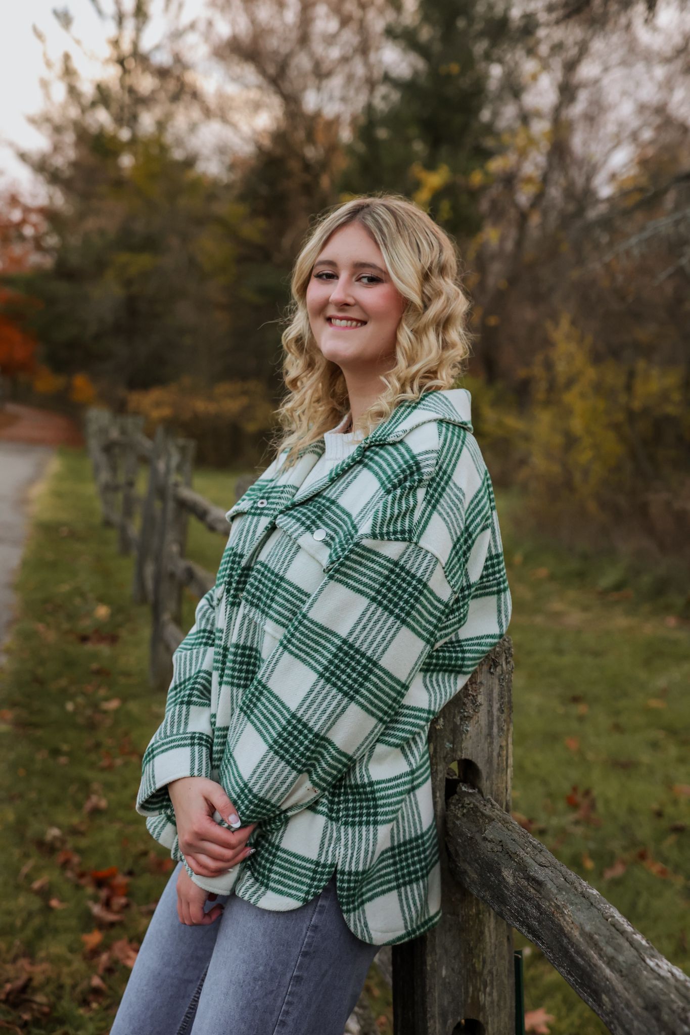 Smiling blonde teen in green plaid flannel jacket leaning on wooden fence in fall foliage.