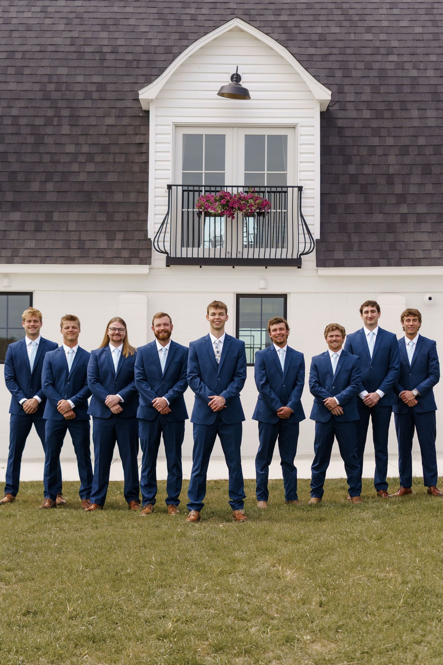 Nine groomsmen in matching navy suits stand in a row outside white farmhouse wedding venue.