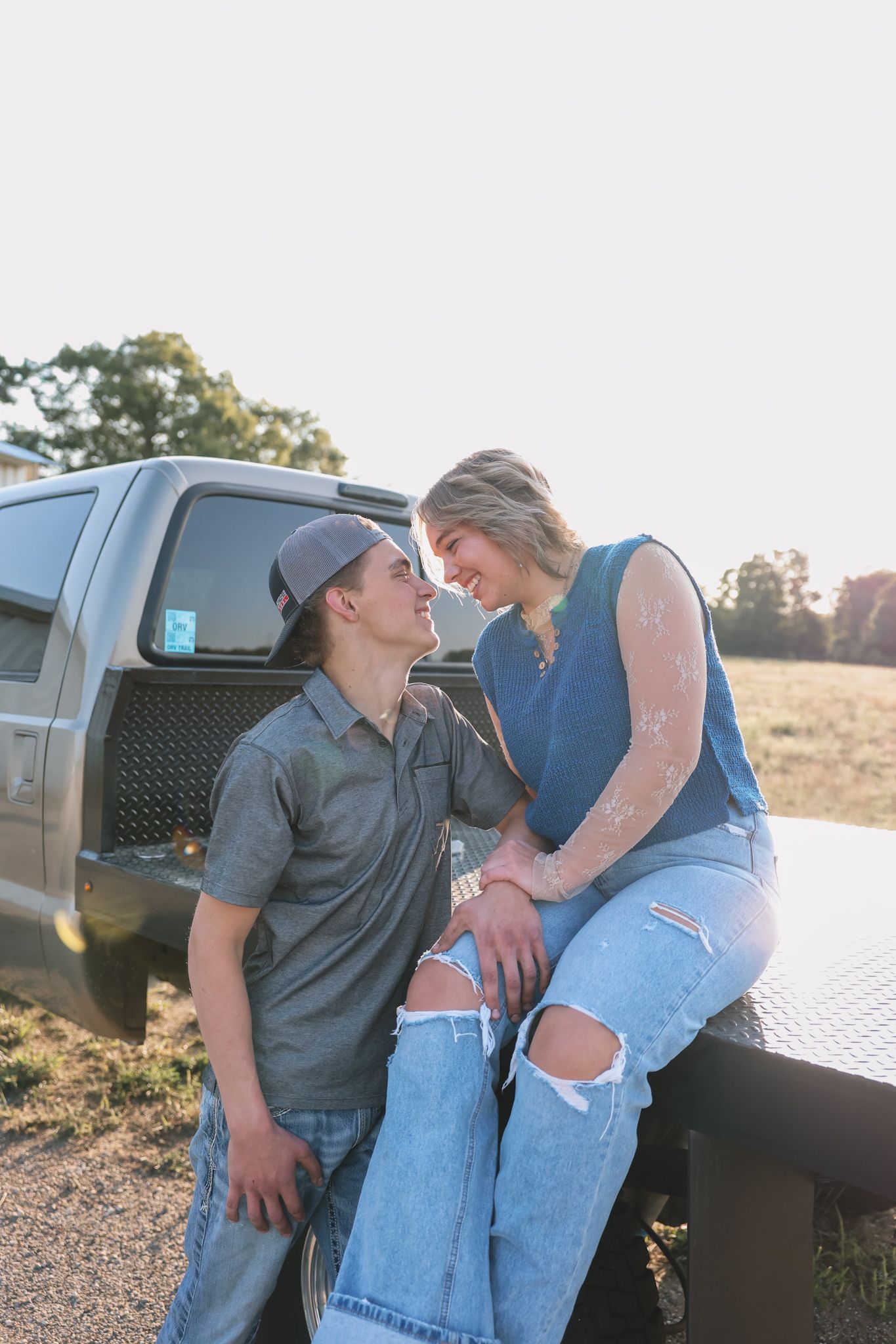 A couple shares a romantic moment on a flatbed truck at golden hour, the woman sitting on the truck bed facing the man.