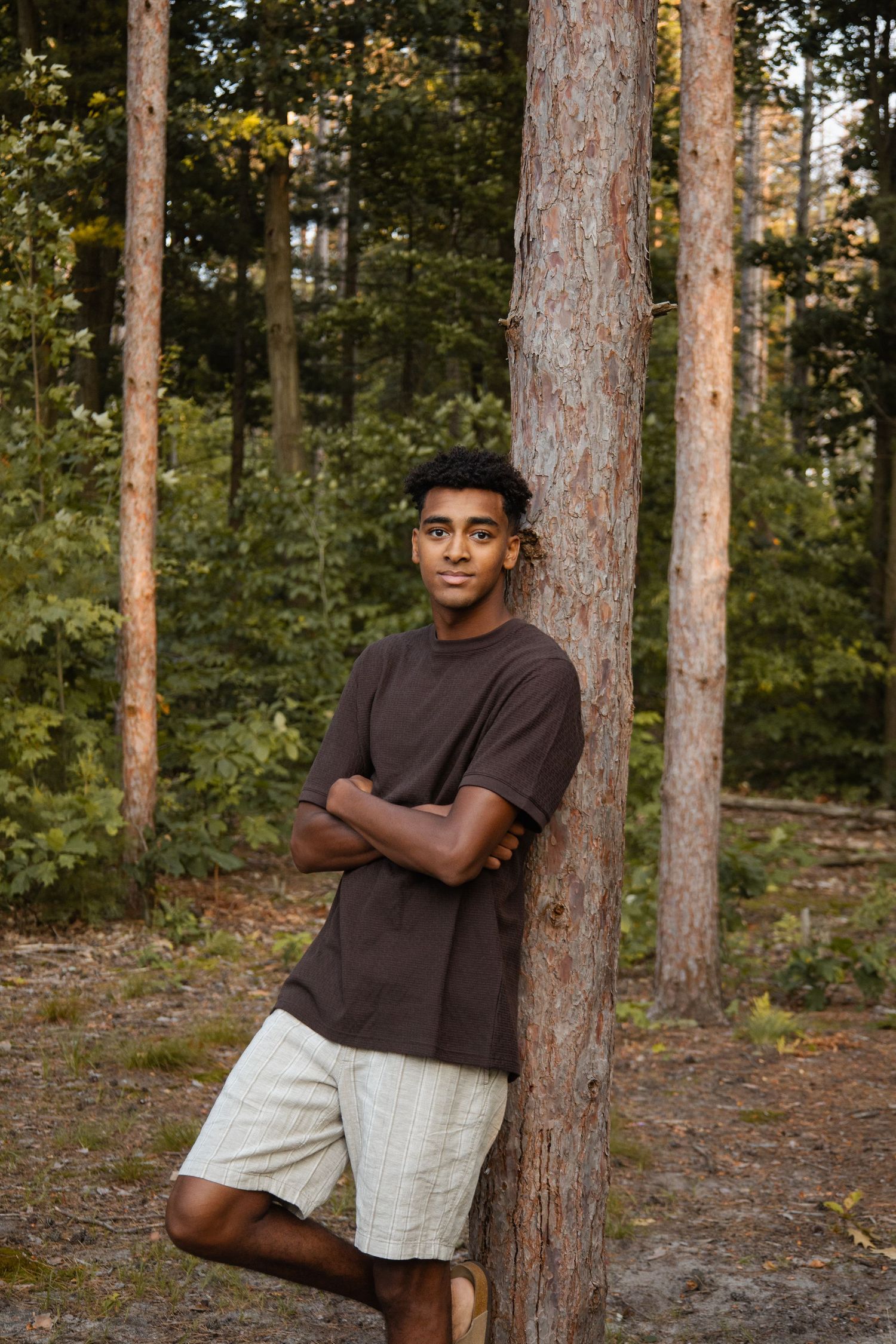 A teenage boy in a brown t-shirt and white shorts leans against a pine tree in a forest setting.
