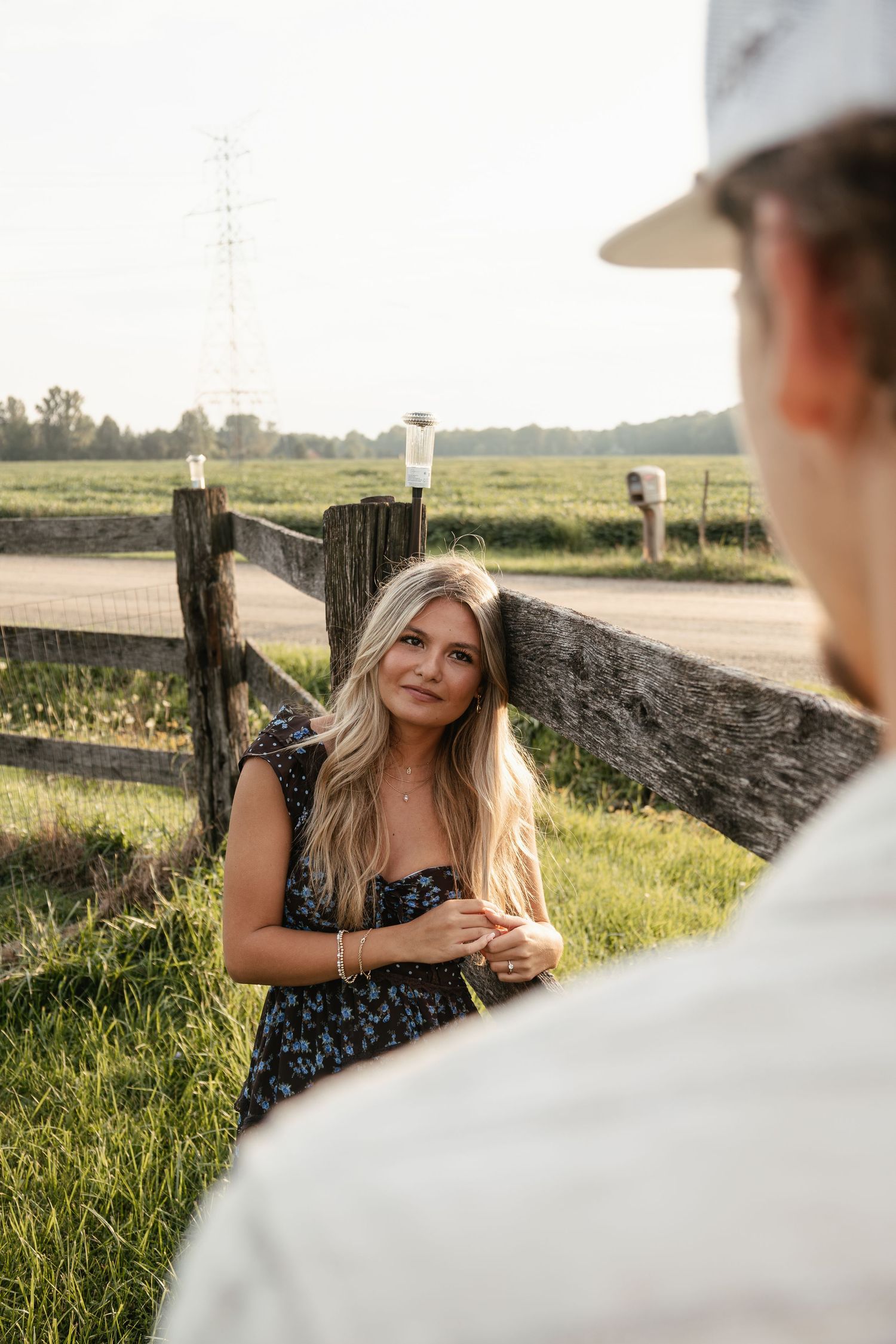 A blonde woman in a floral dress leans against a wooden fence in a rural field, smiling at someone in the foreground.