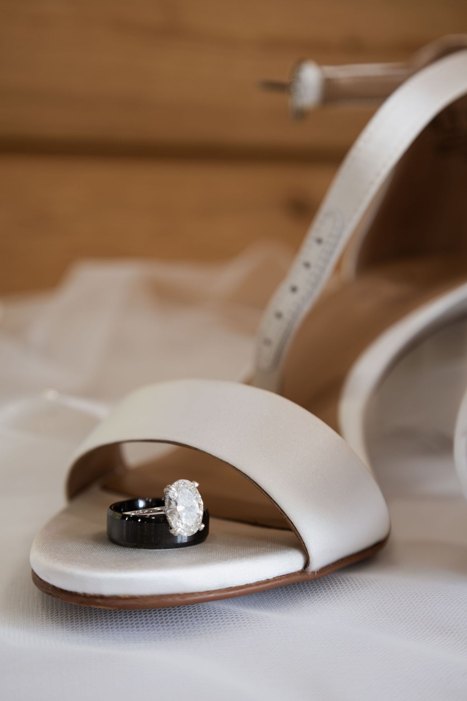 Wedding rings resting in a white bridal heel with diamond engagement ring and black band on white veil.