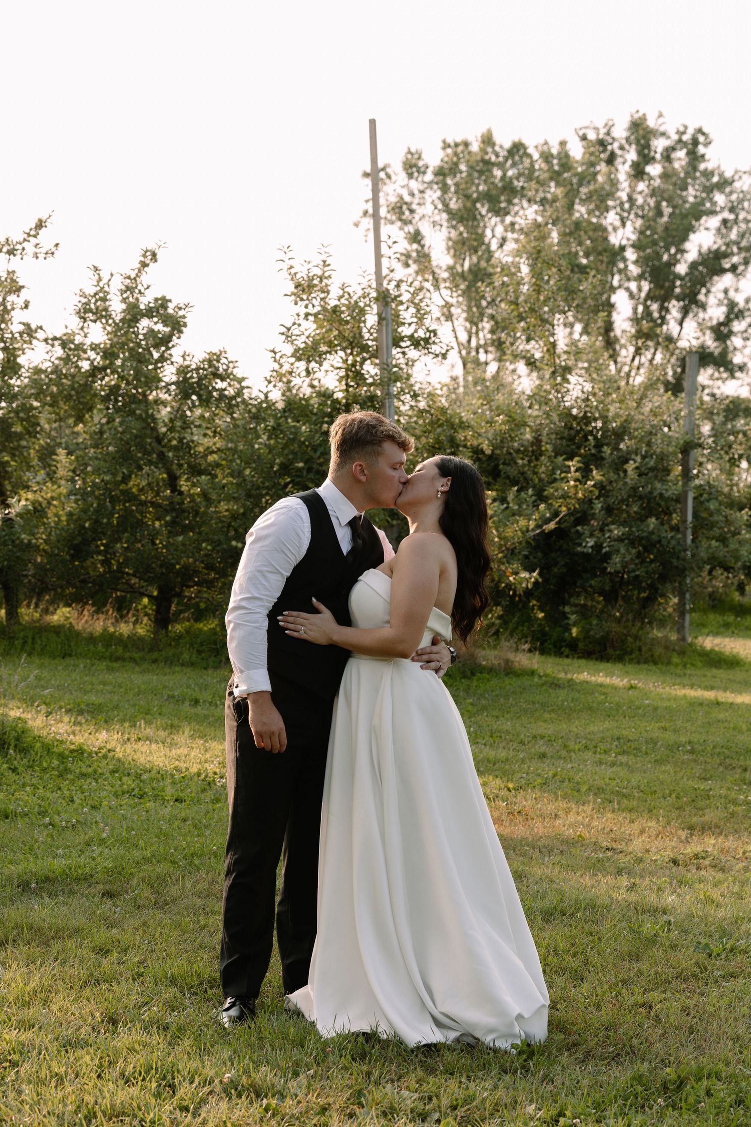 Bride and groom share a kiss in golden hour light surrounded by lush green orchard.
