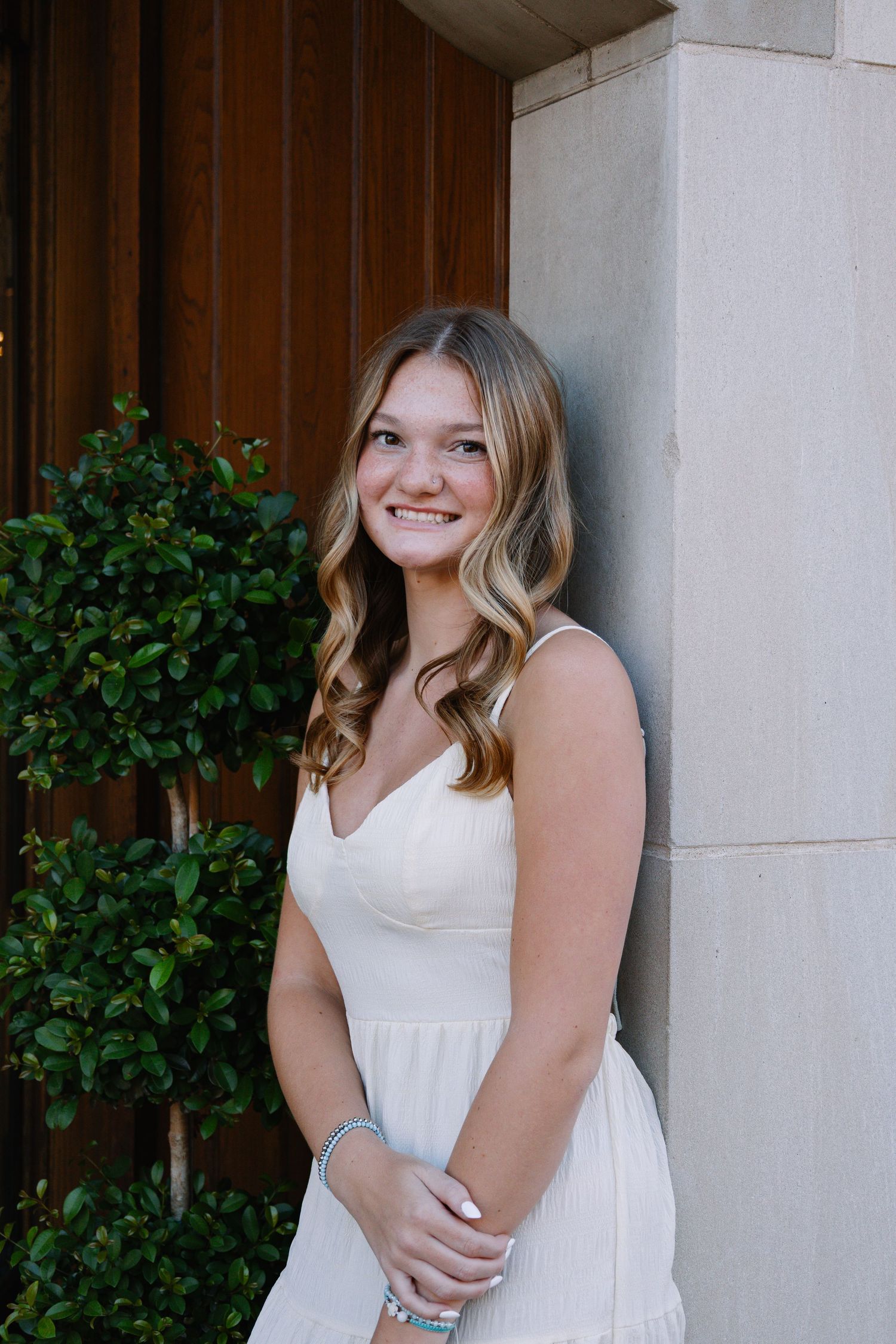 Young woman in white dress smiling outdoors near stone column and green shrubs.
