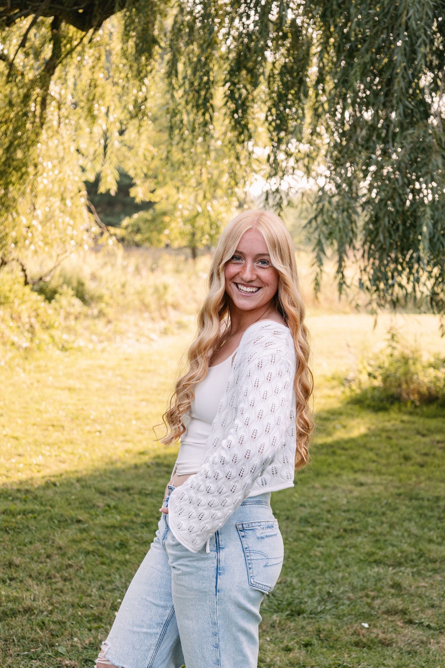 Smiling blonde woman in white lace top and jeans poses outdoors under a willow tree in golden sunlight.