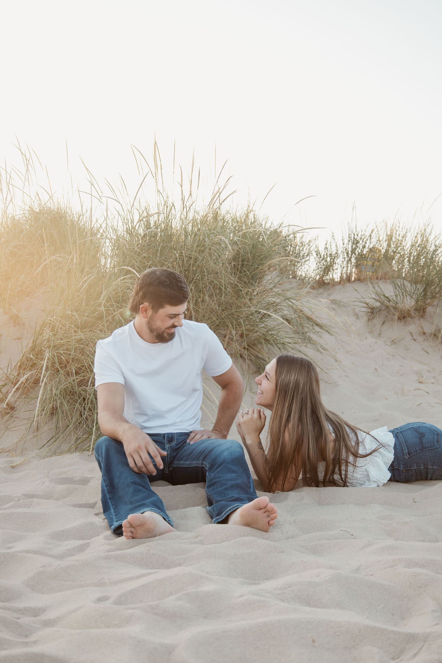 Couple sitting on sandy beach dunes, woman lying down facing man in white tee and jeans, golden hour light.