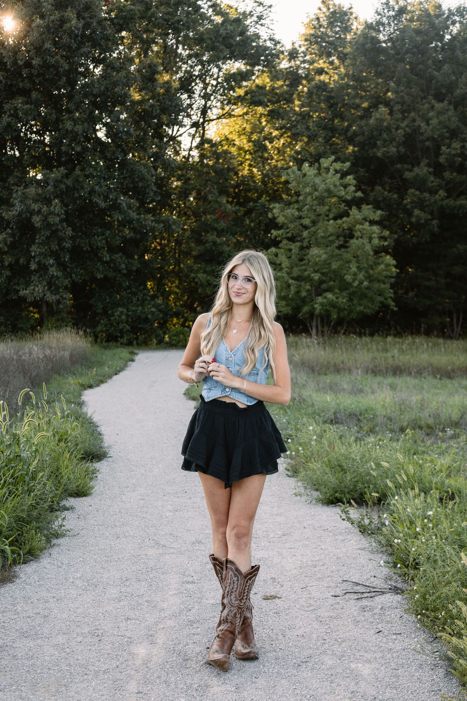 Blonde woman in blue crop top, black mini skirt, and cowboy boots standing on a gravel path surrounded by trees.
