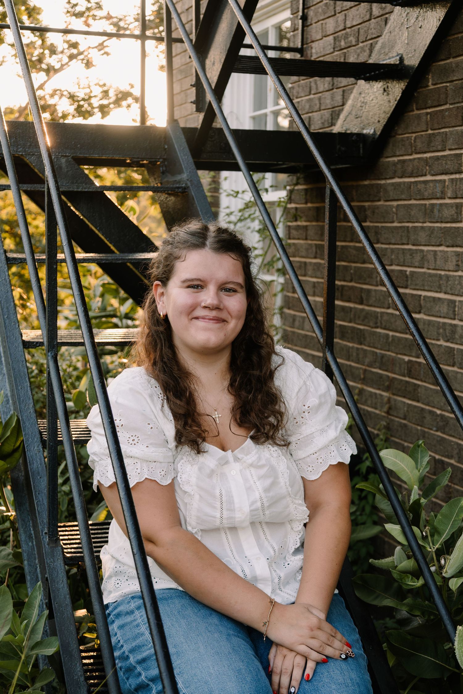 Young woman in white eyelet top and jeans smiling on metal spiral staircase near brick wall and greenery.