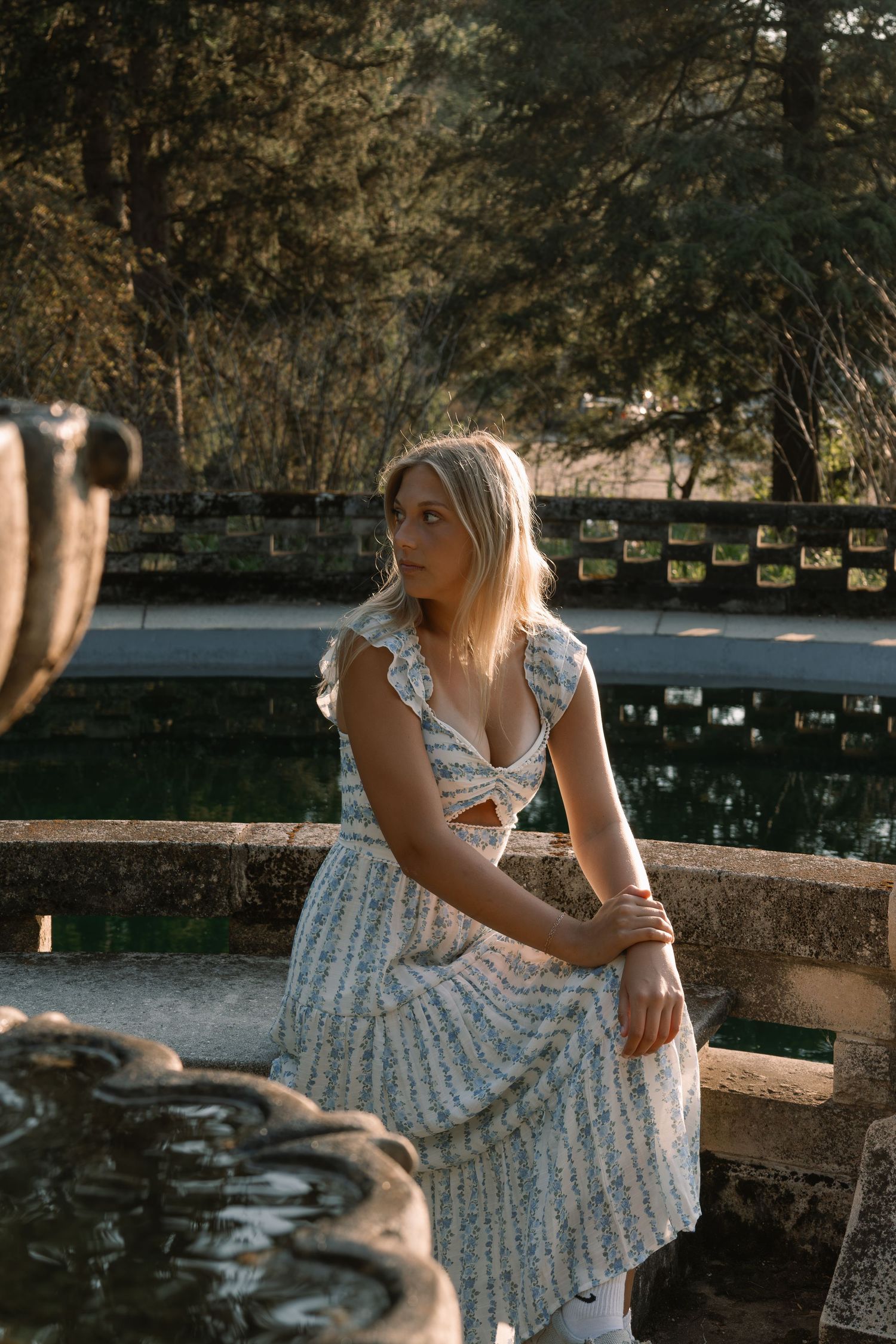 Young blonde woman in floral sundress sitting on stone fountain edge in sunlit garden park.