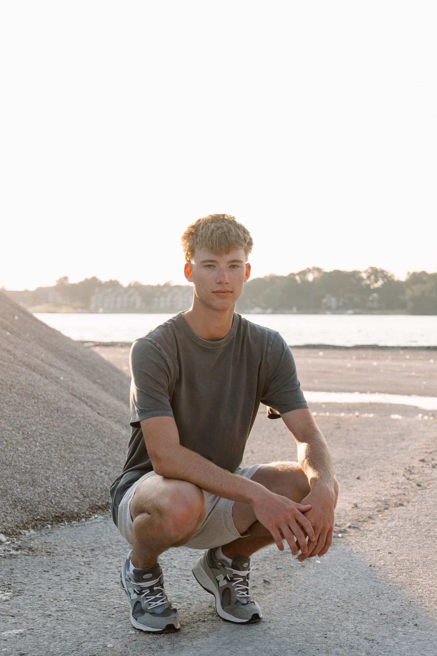 Young man in gray t-shirt and shorts crouching on sandy beach path at golden hour sunset.