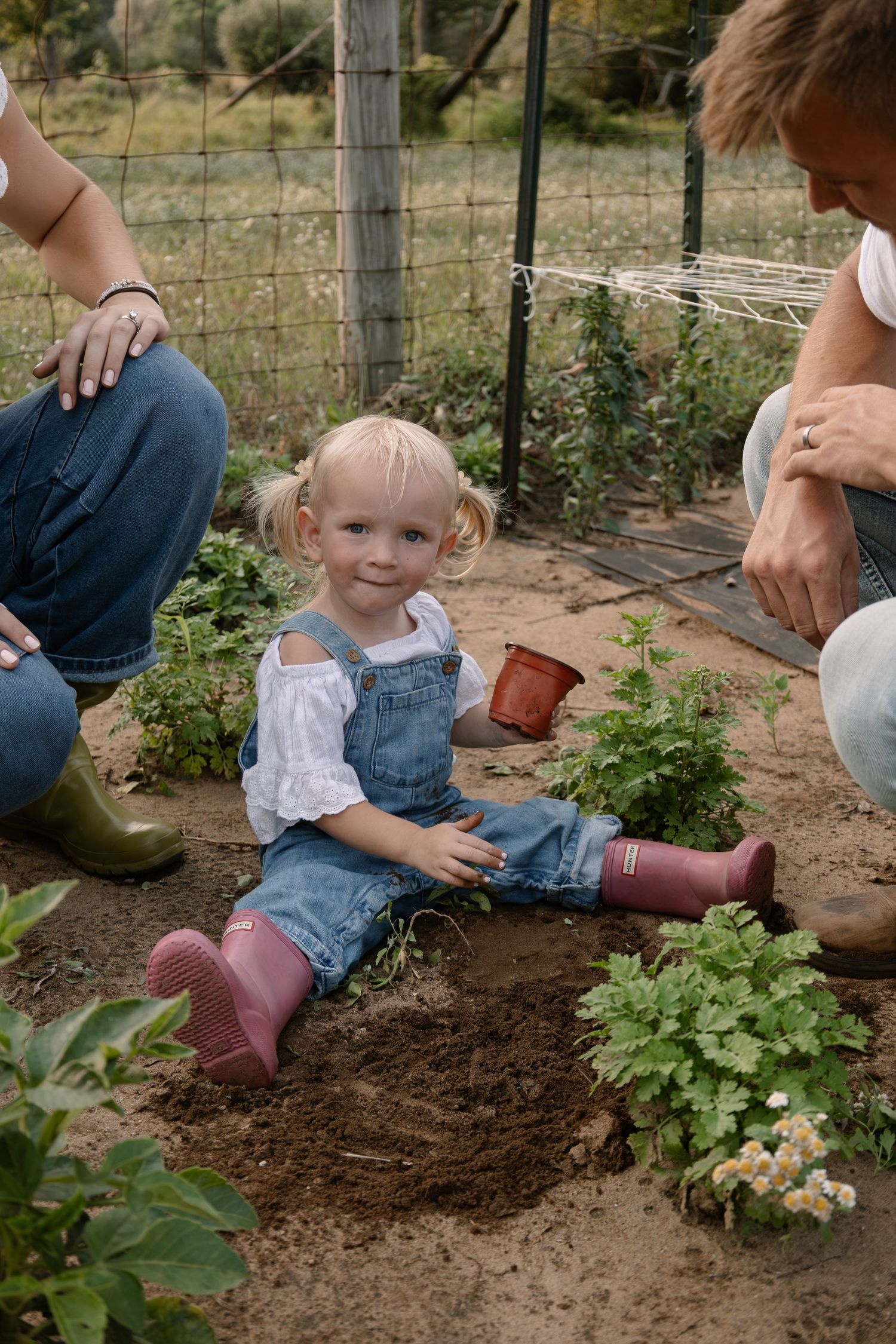 Toddler girl in denim overalls and pink boots sits in garden dirt while adults garden nearby.