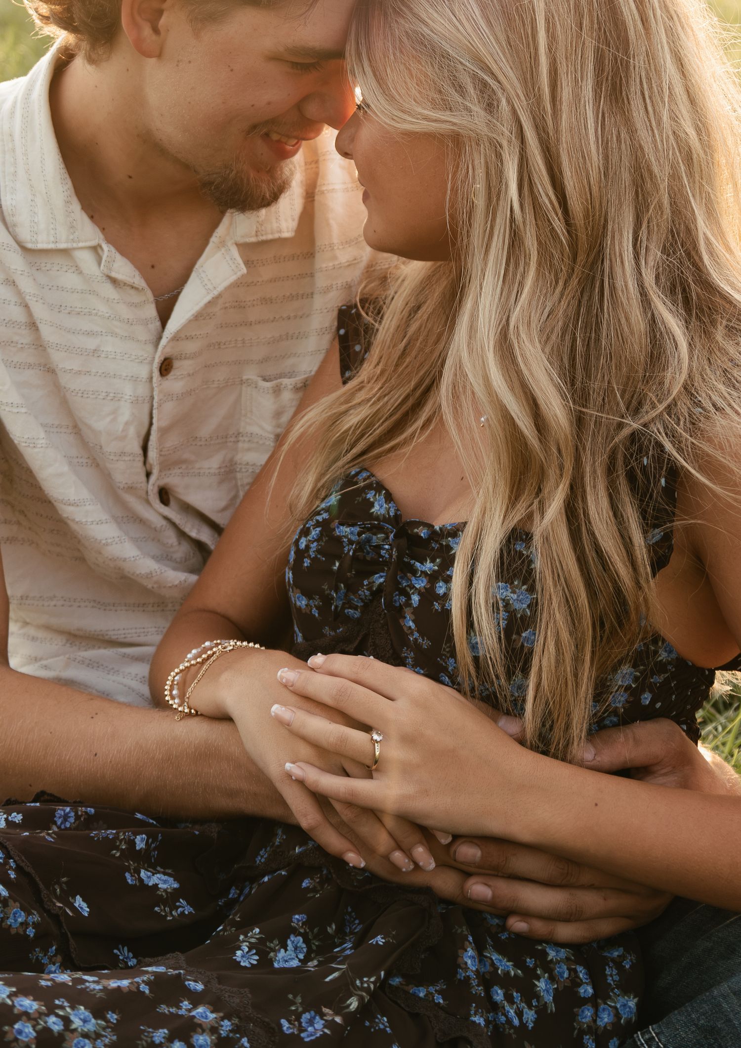 Couple in golden light, woman in floral dress showing engagement ring, man in striped shirt embracing her tenderly.
