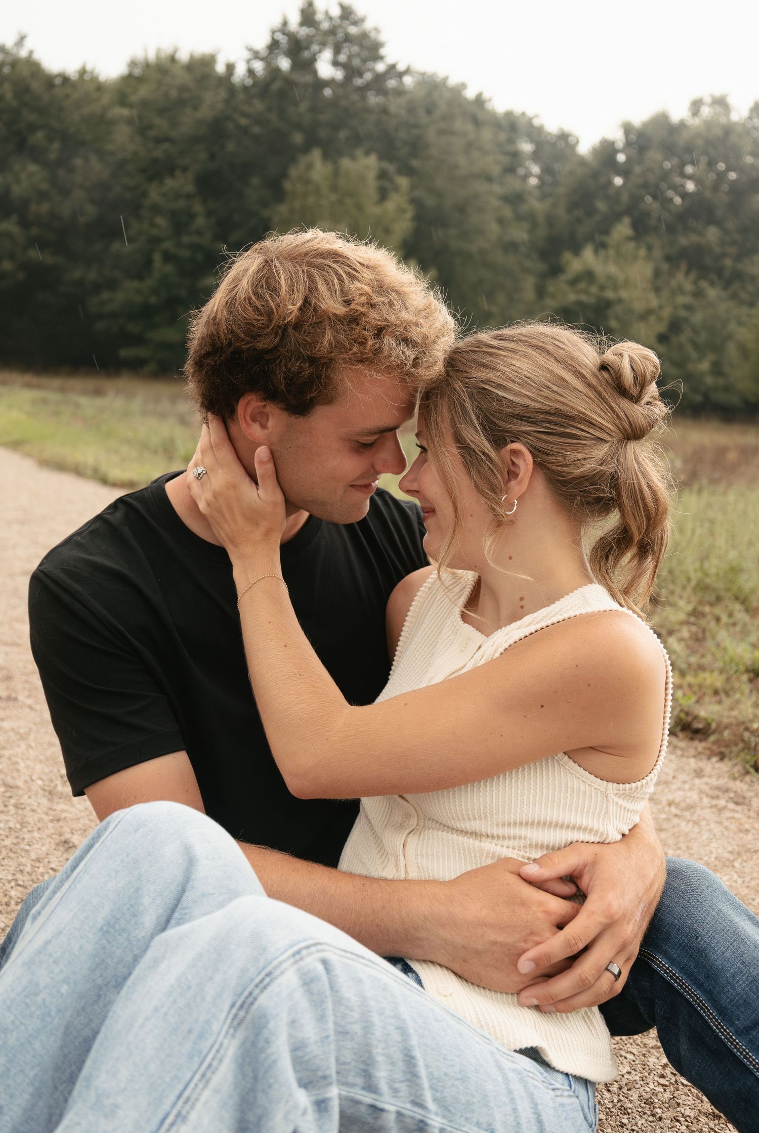 A young couple sits closely on a dirt path, sharing a tender moment surrounded by lush green trees.
