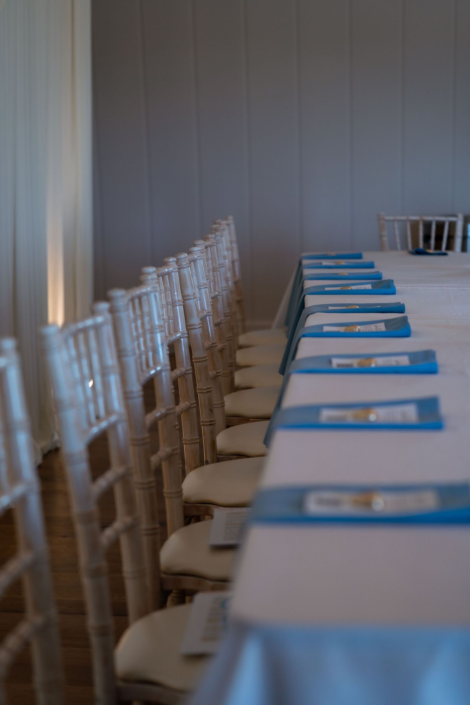 Long banquet table with white linens, blue napkins, and chiavari chairs in elegant wedding venue.