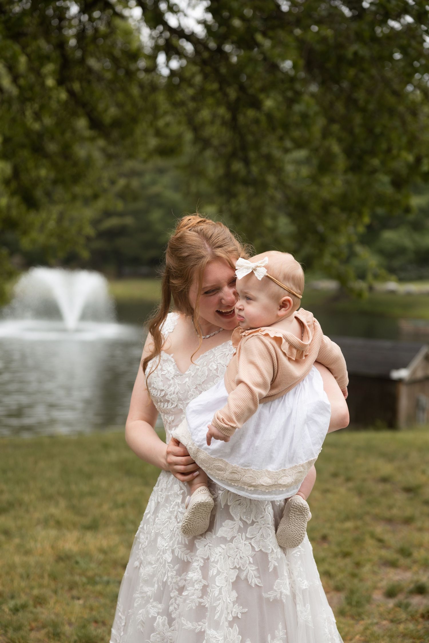 Bride in lace wedding gown holding a baby girl in white dress near a garden fountain.