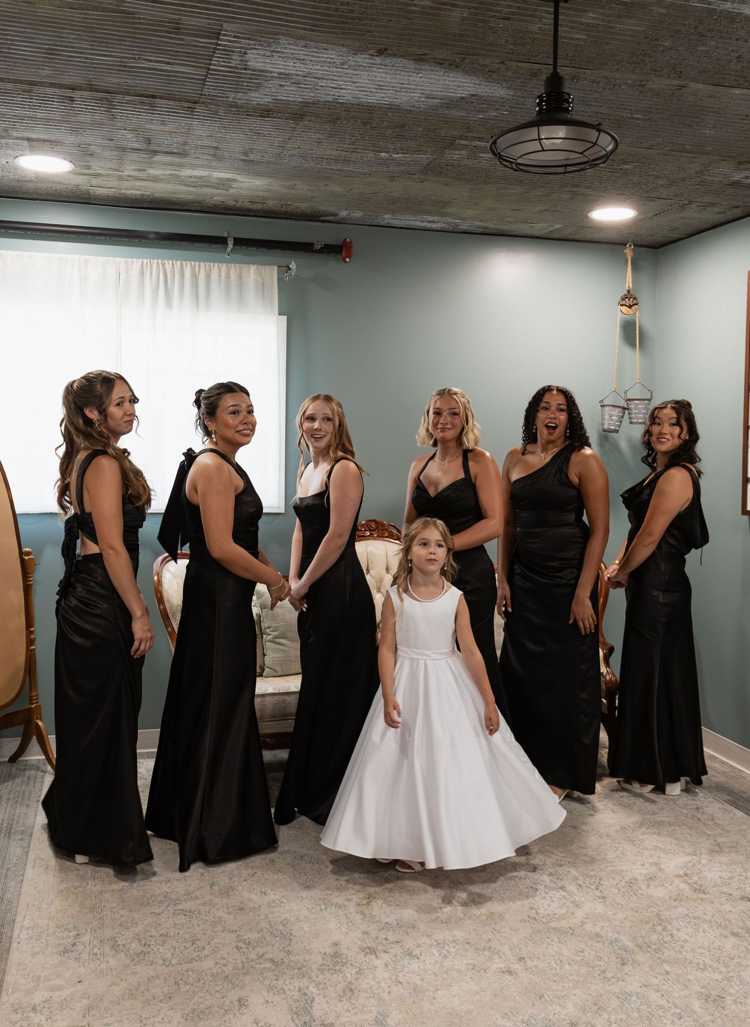 Bridesmaids in black gowns pose with a flower girl in white dress indoors at a wedding venue.