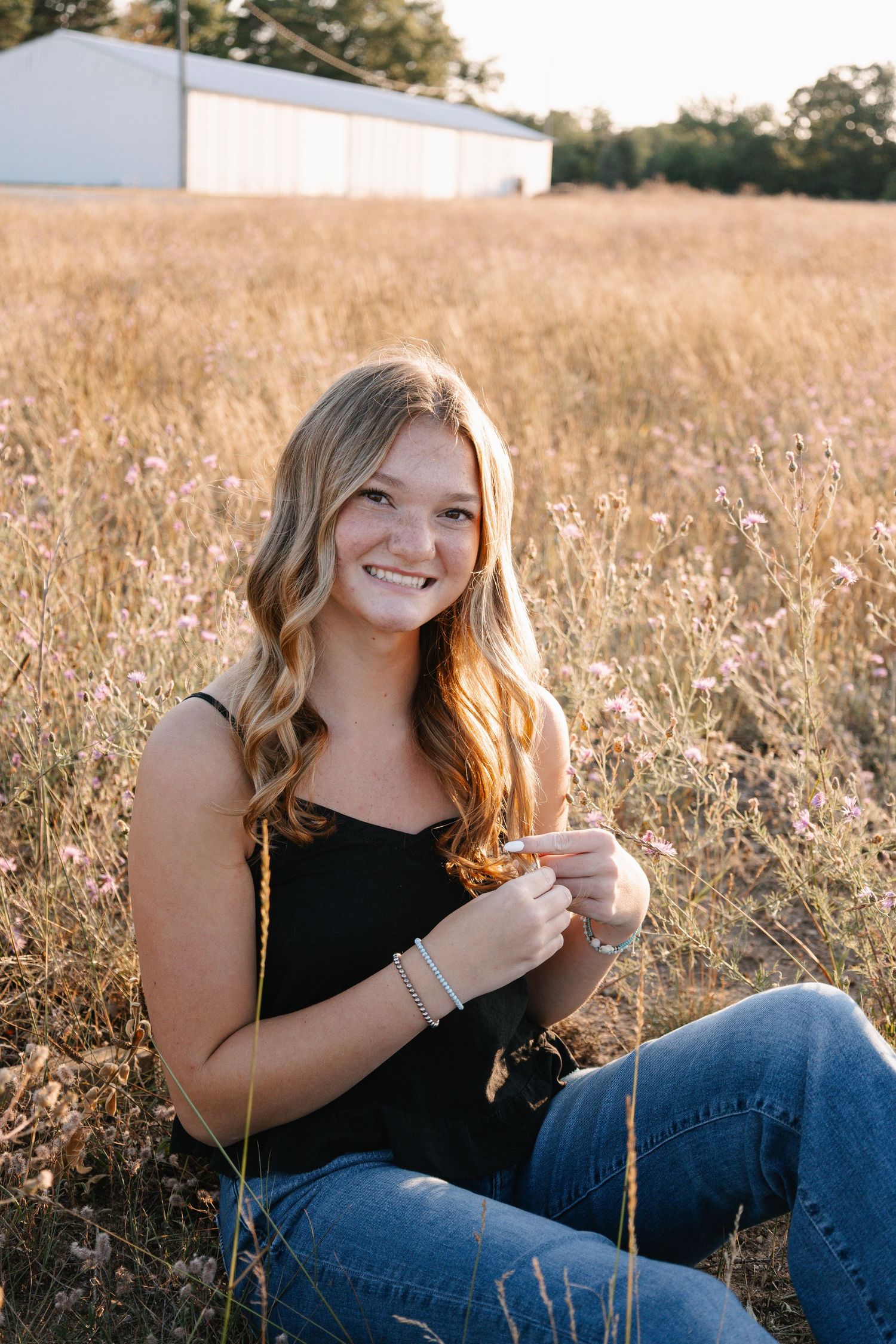 Young woman with wavy blonde hair sitting in golden field, smiling, wearing black tank top and jeans.