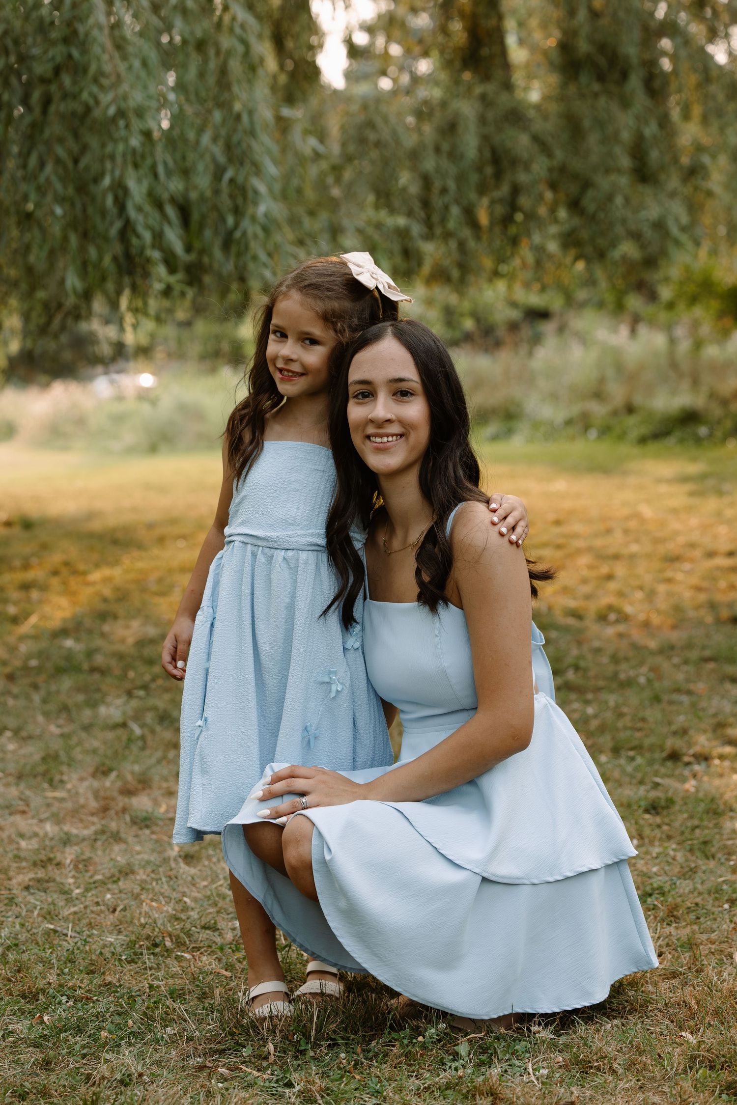 A woman and young girl in matching light blue dresses pose together outdoors in a lush green park setting.