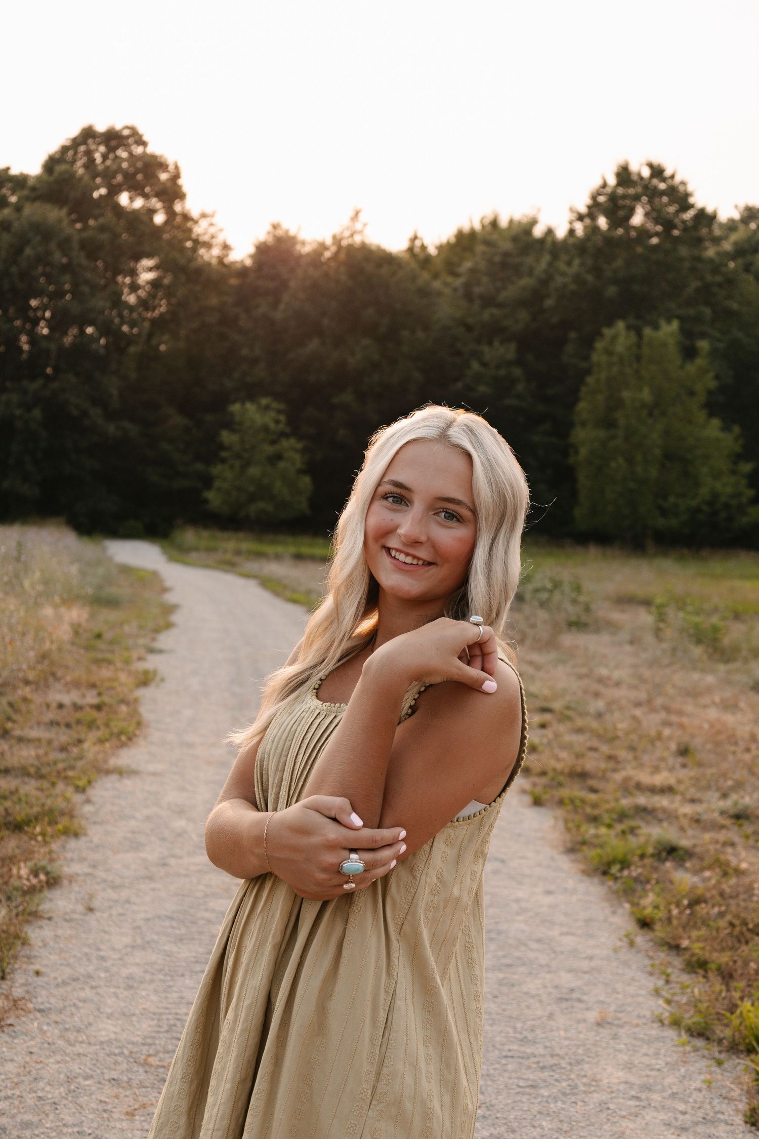 Smiling blonde woman in olive dress poses on dirt path at golden hour with green trees in background.
