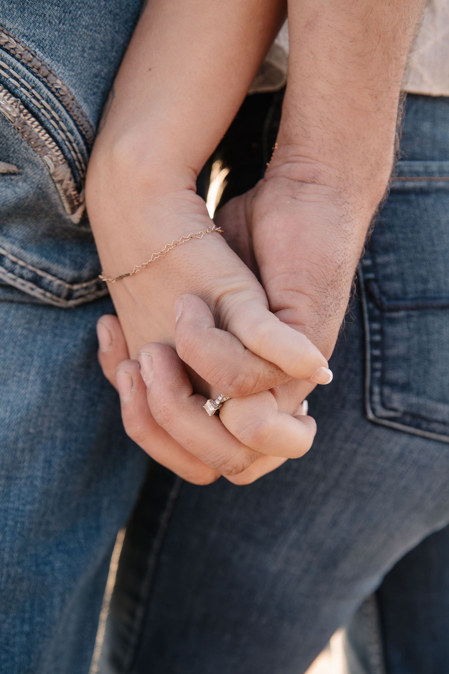 Close-up of a couple holding hands, woman wearing a diamond engagement ring and gold bracelet, both in jeans.