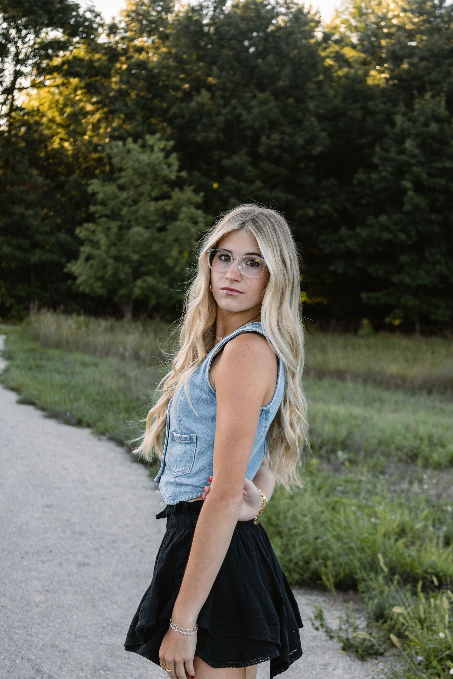 Young blonde woman in denim vest and black skirt posing on a path surrounded by lush green trees.
