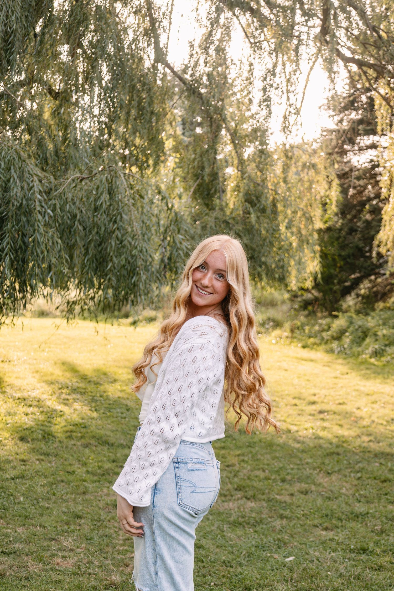 Young woman with long blonde wavy hair smiling outdoors in white top and jeans near a willow tree.