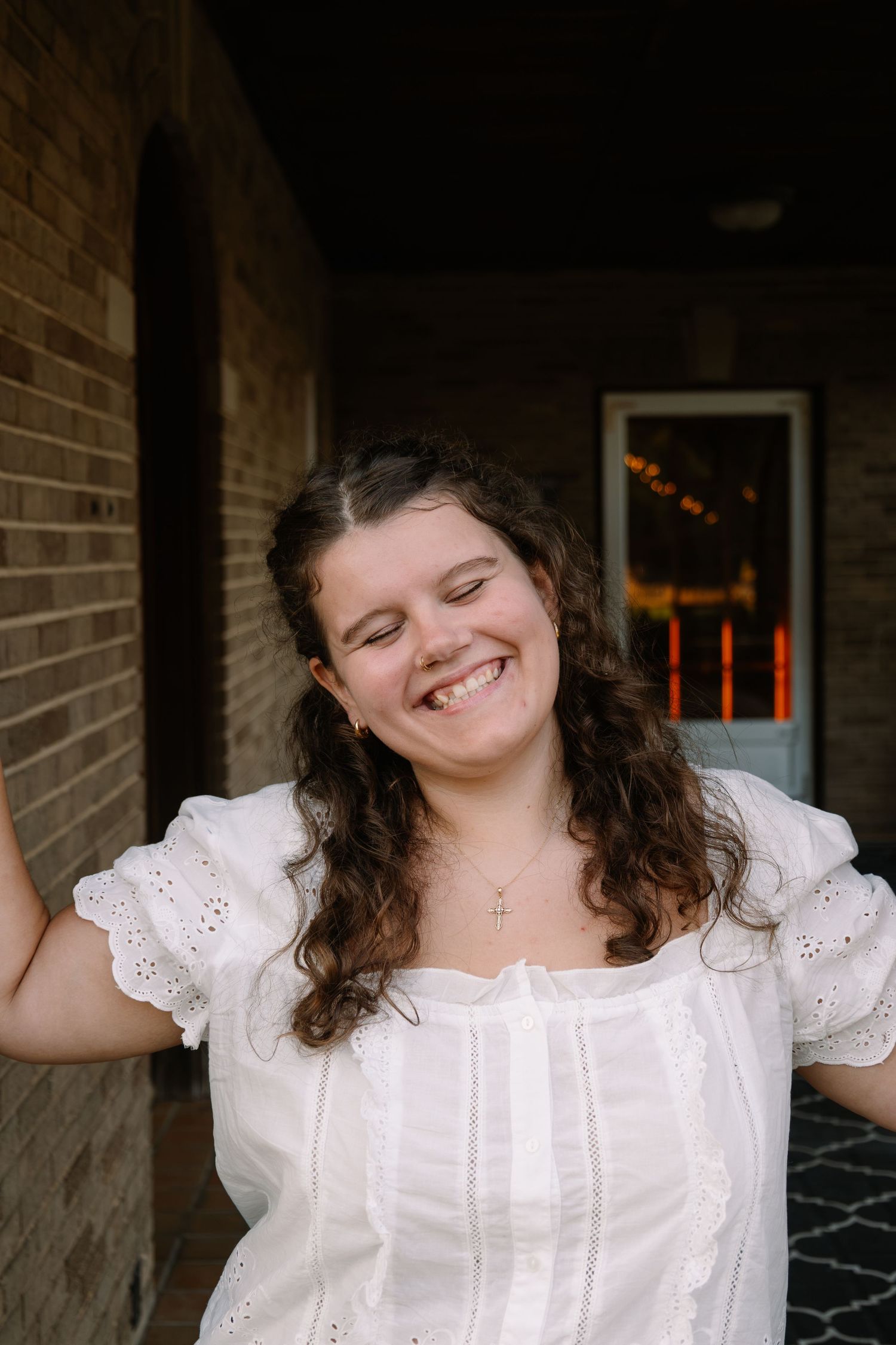 Young woman in white eyelet dress laughing joyfully outdoors near brick wall.
