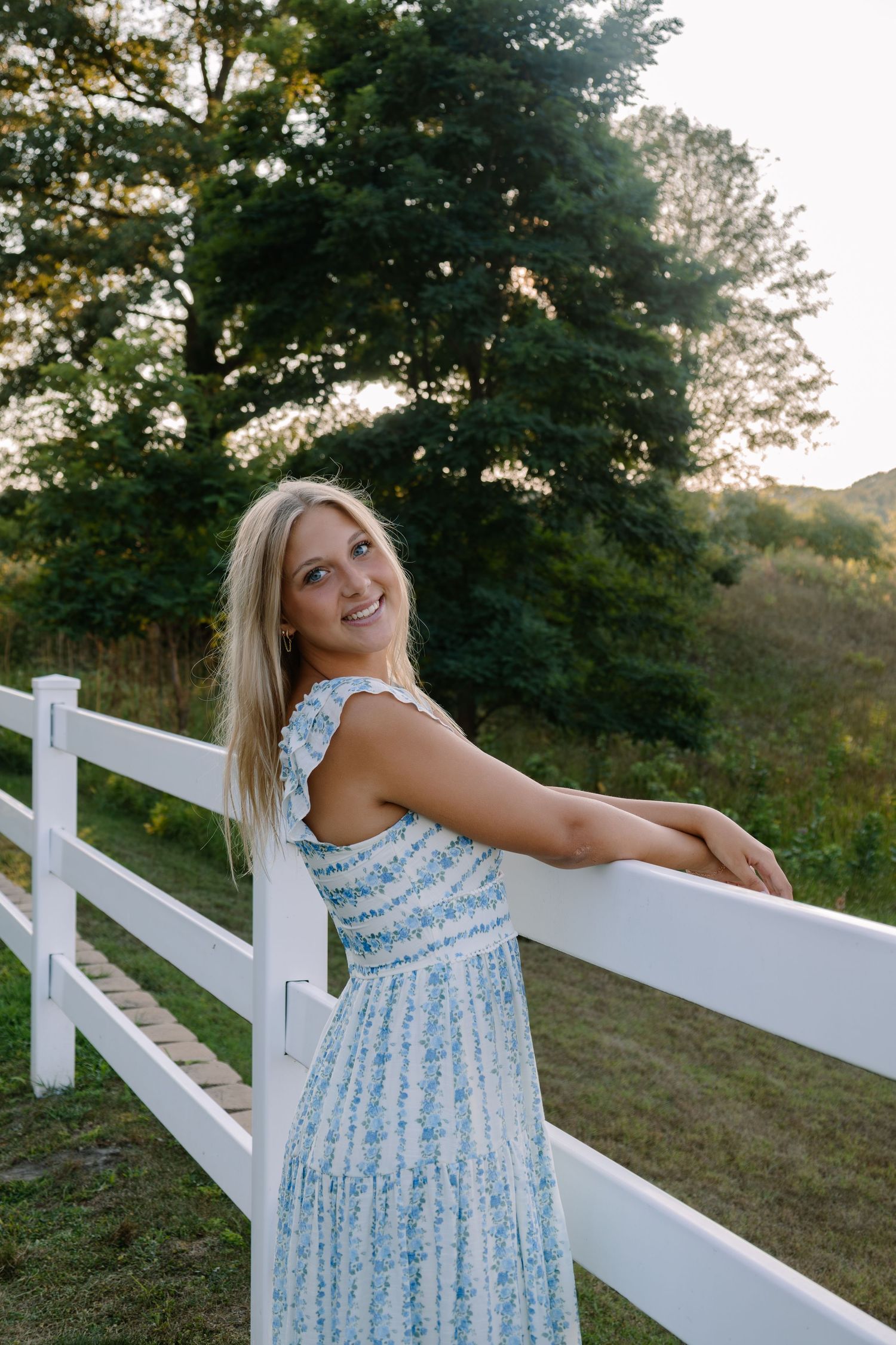 Smiling blonde woman in a blue floral dress leaning on a white fence with lush green trees in the background.