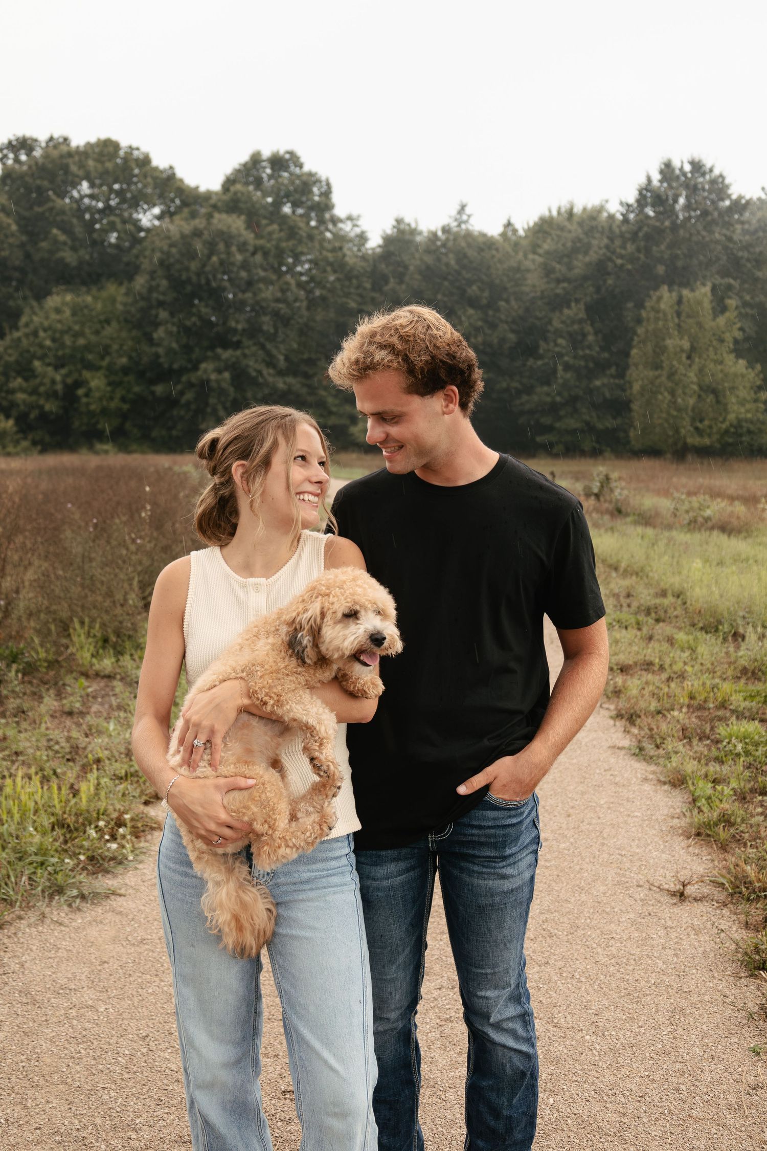 A smiling couple holds a fluffy golden doodle puppy on a scenic dirt path surrounded by lush greenery.