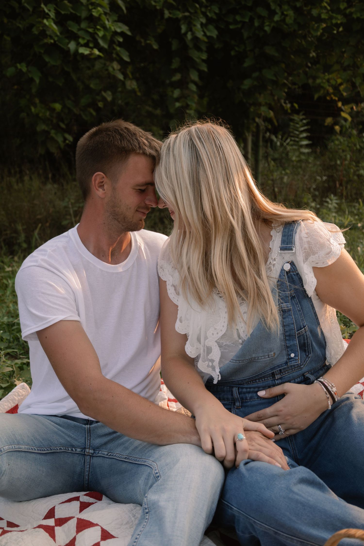 A couple sitting on a quilt outdoors, touching foreheads romantically, woman in denim overalls and man in white tee.