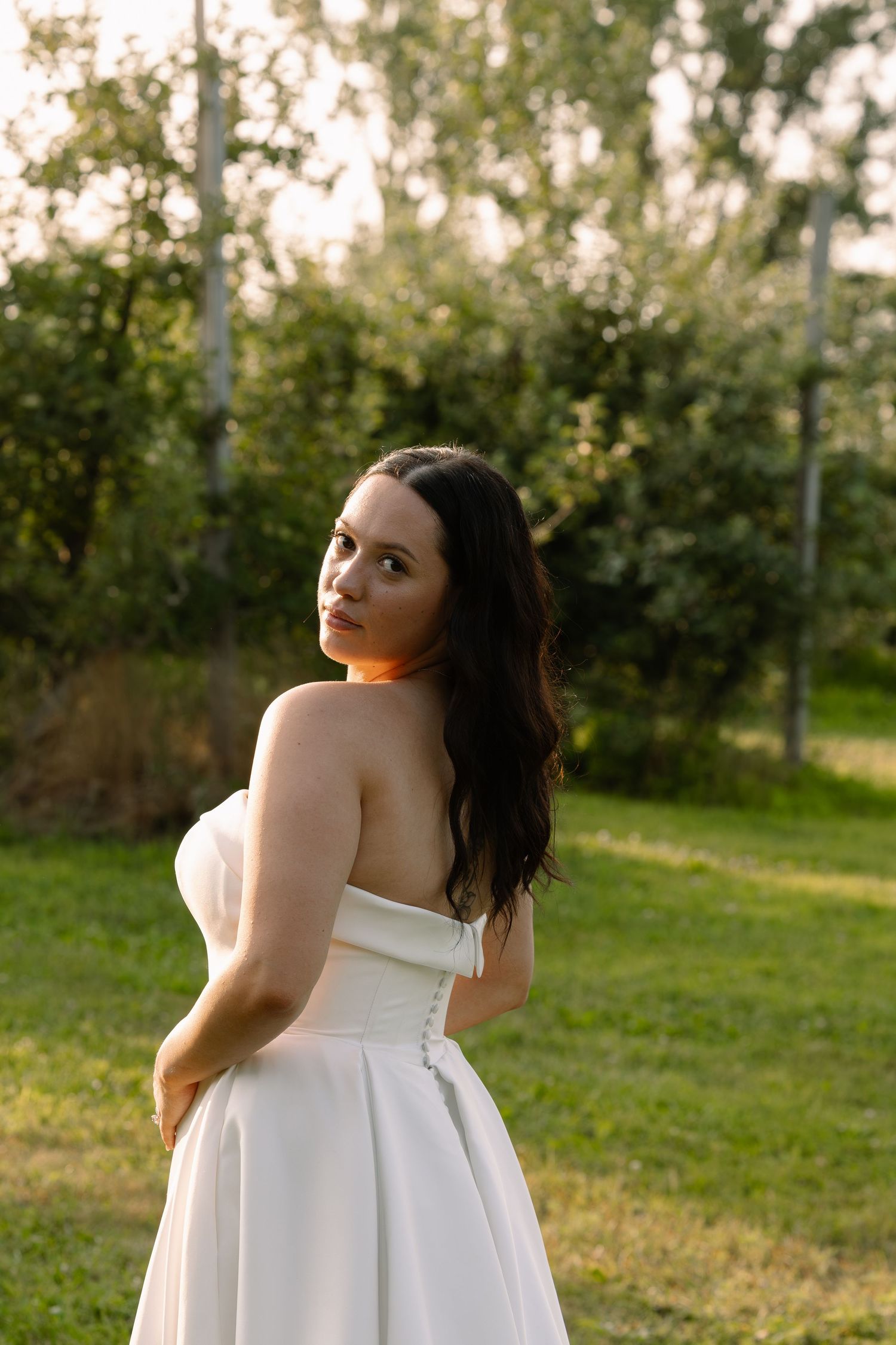 Brunette bride in strapless white gown poses outdoors in lush green garden at golden hour.