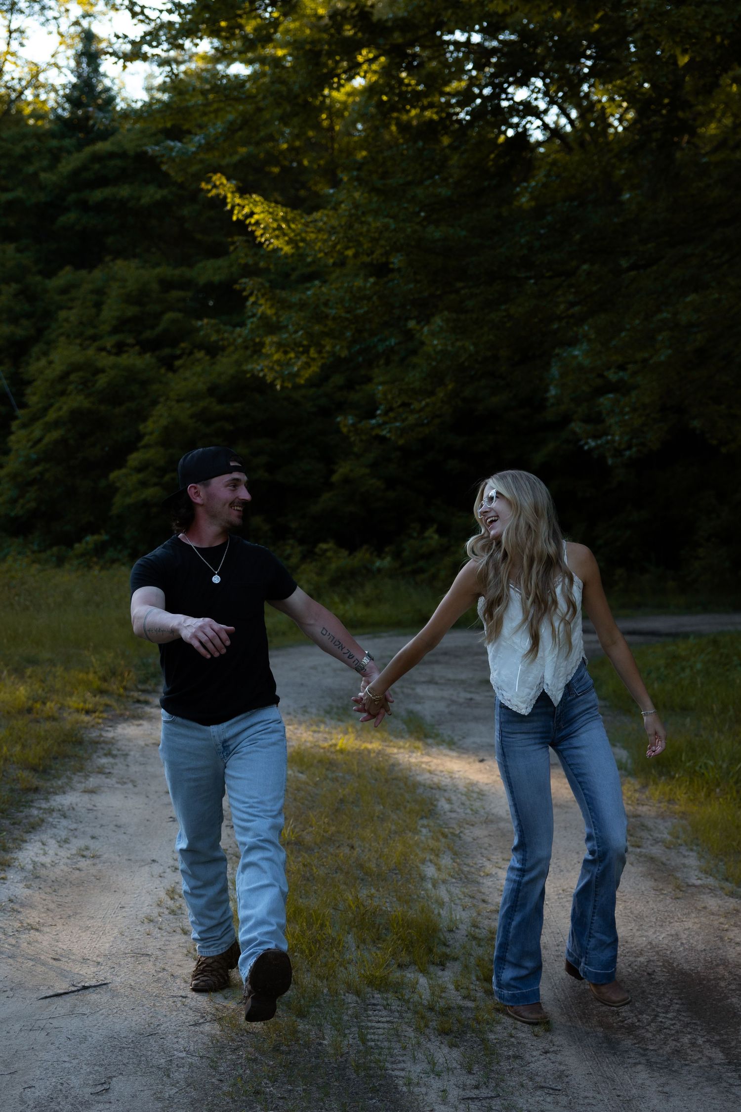 Couple holding hands and laughing while walking down a rural dirt path surrounded by lush green trees at dusk.