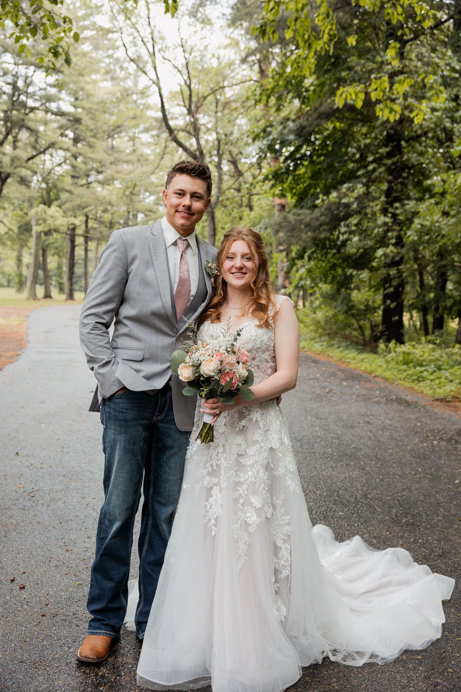 Bride and groom posing together on a tree-lined path, bride holding a pink floral bouquet.