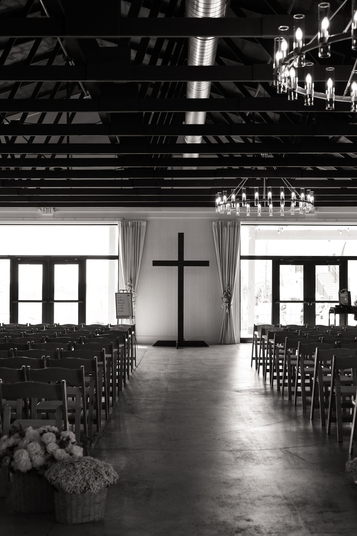 Black and white photo of a rustic wedding chapel interior with wooden cross, chandelier, and rows of chairs.