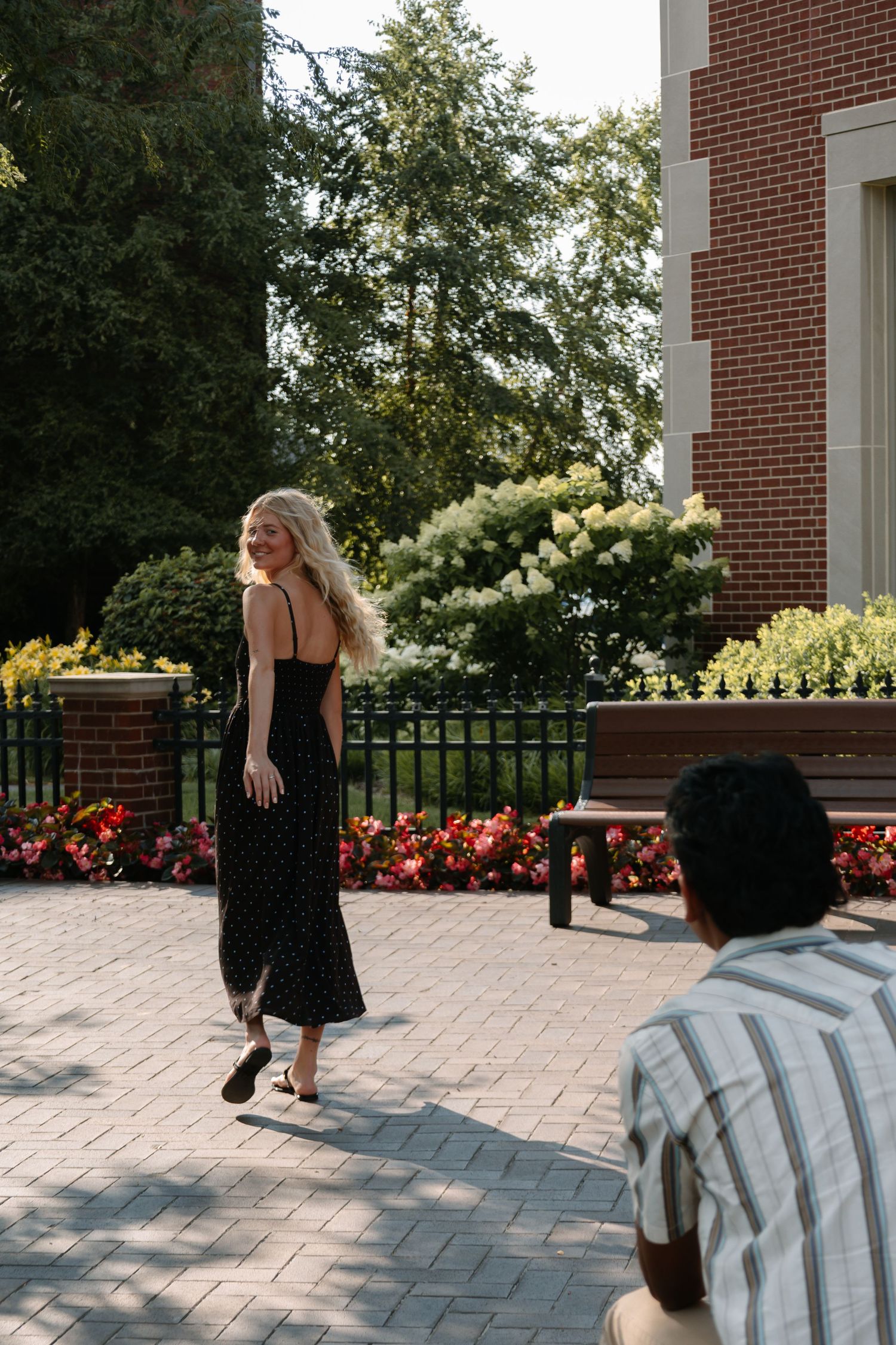 A blonde woman in a black floral midi dress walks on a brick patio while a man watches near a garden.
