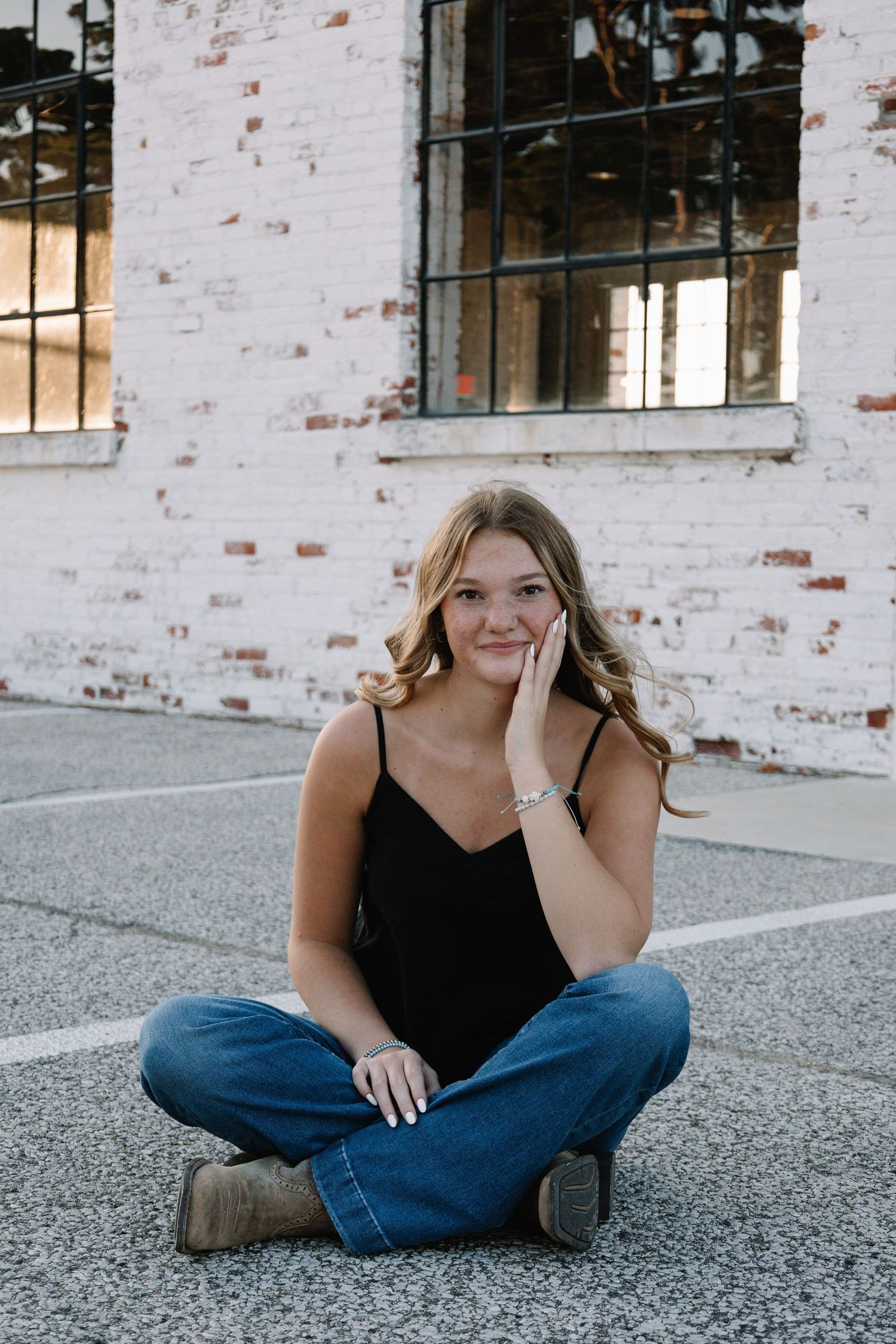 Young blonde woman sitting cross-legged on pavement in black tank and jeans, posing near brick building.