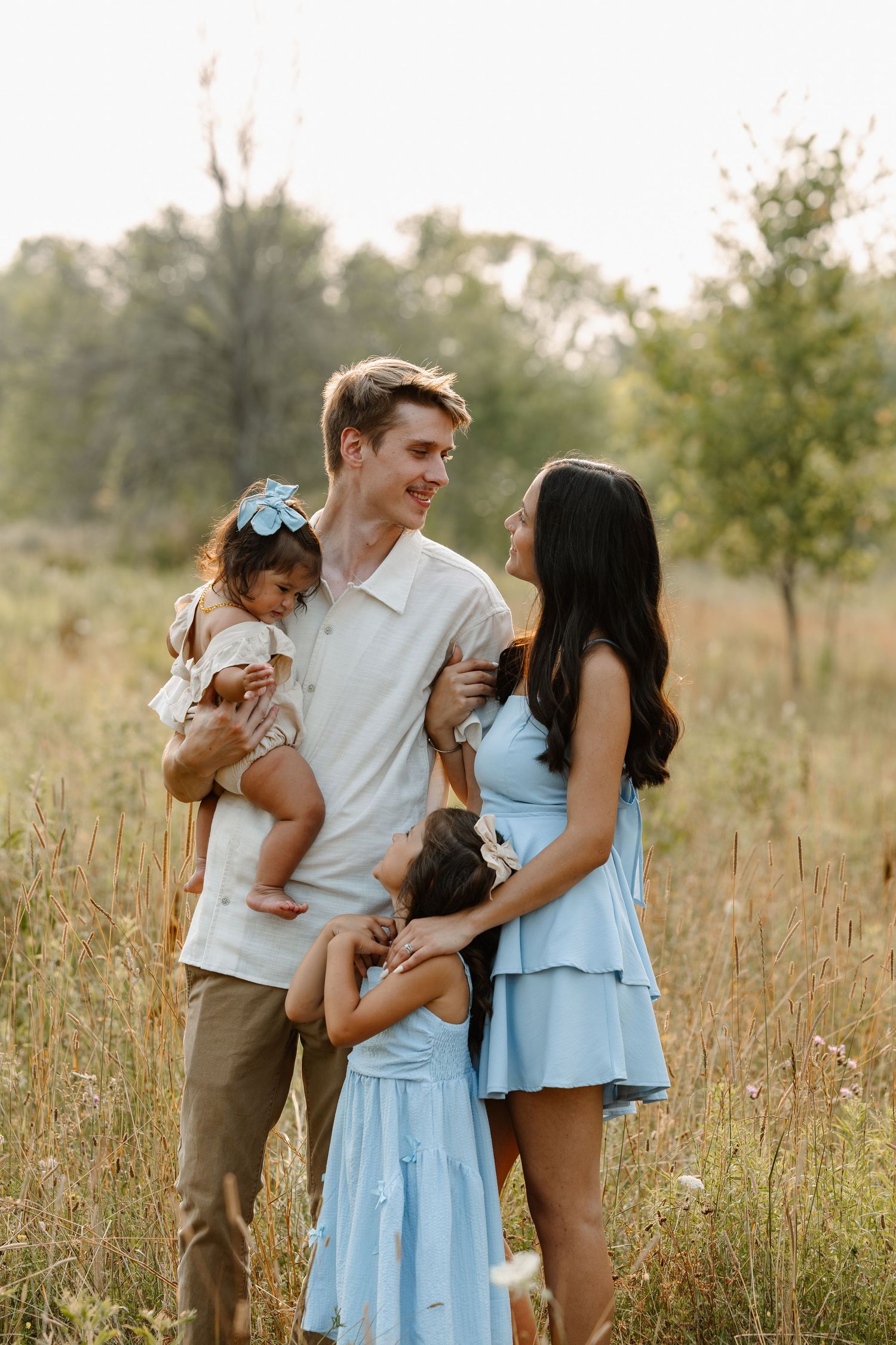 A happy young family poses in a sunlit meadow, with a man holding a baby and a woman in a blue dress hugging their toddler.