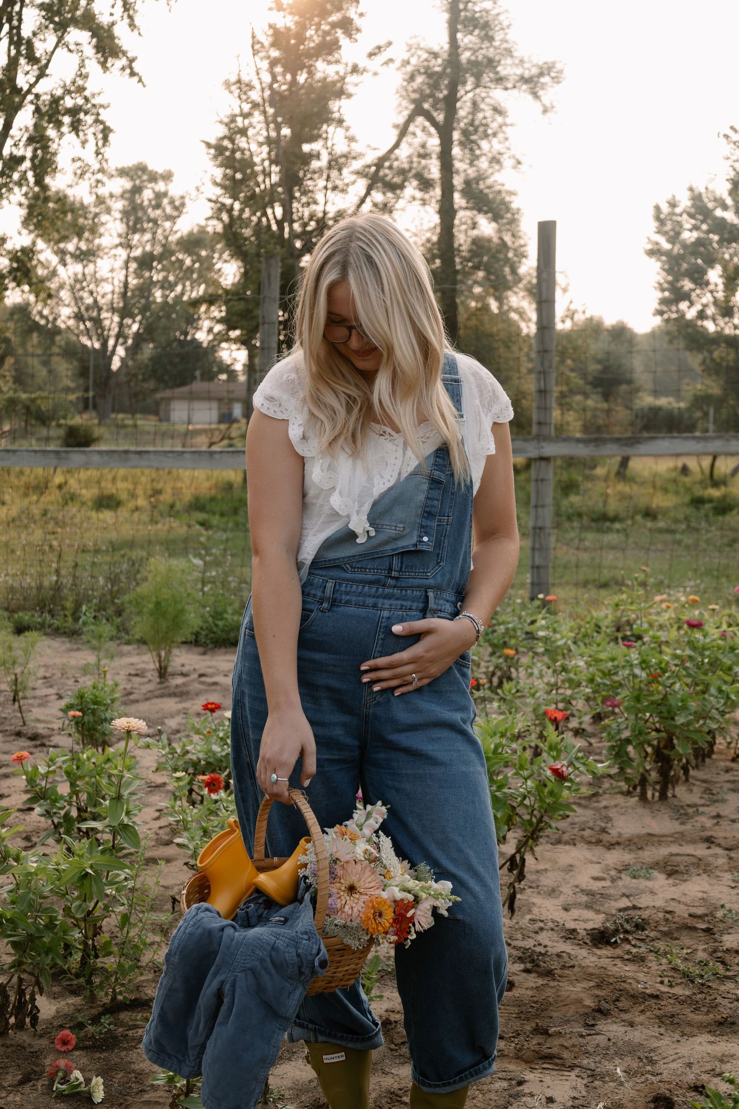 Blonde pregnant woman in denim overalls holding a flower basket while standing in a sunlit flower garden.