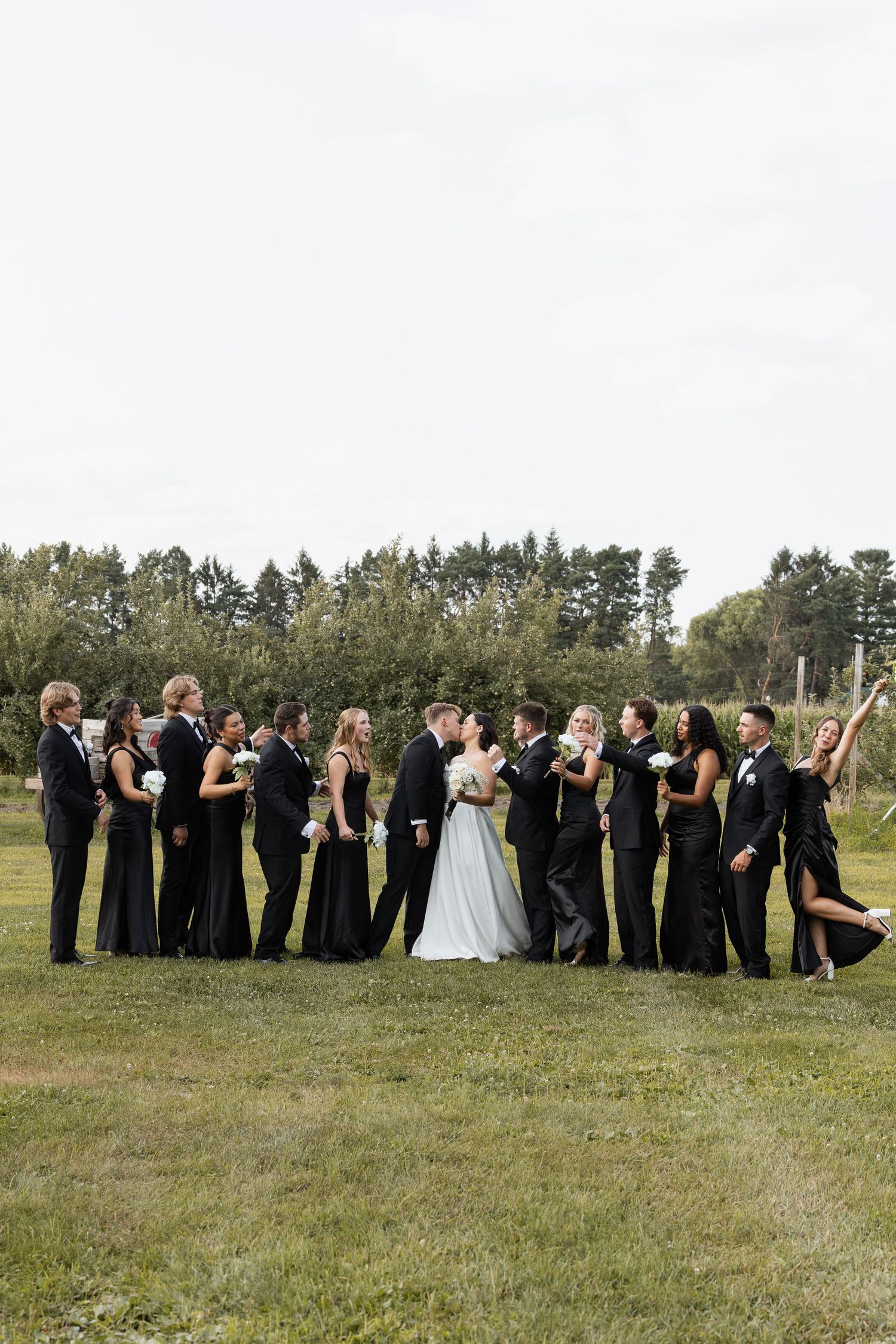 Wedding party in black attire with bride and groom sharing a kiss on a green lawn with trees in background.
