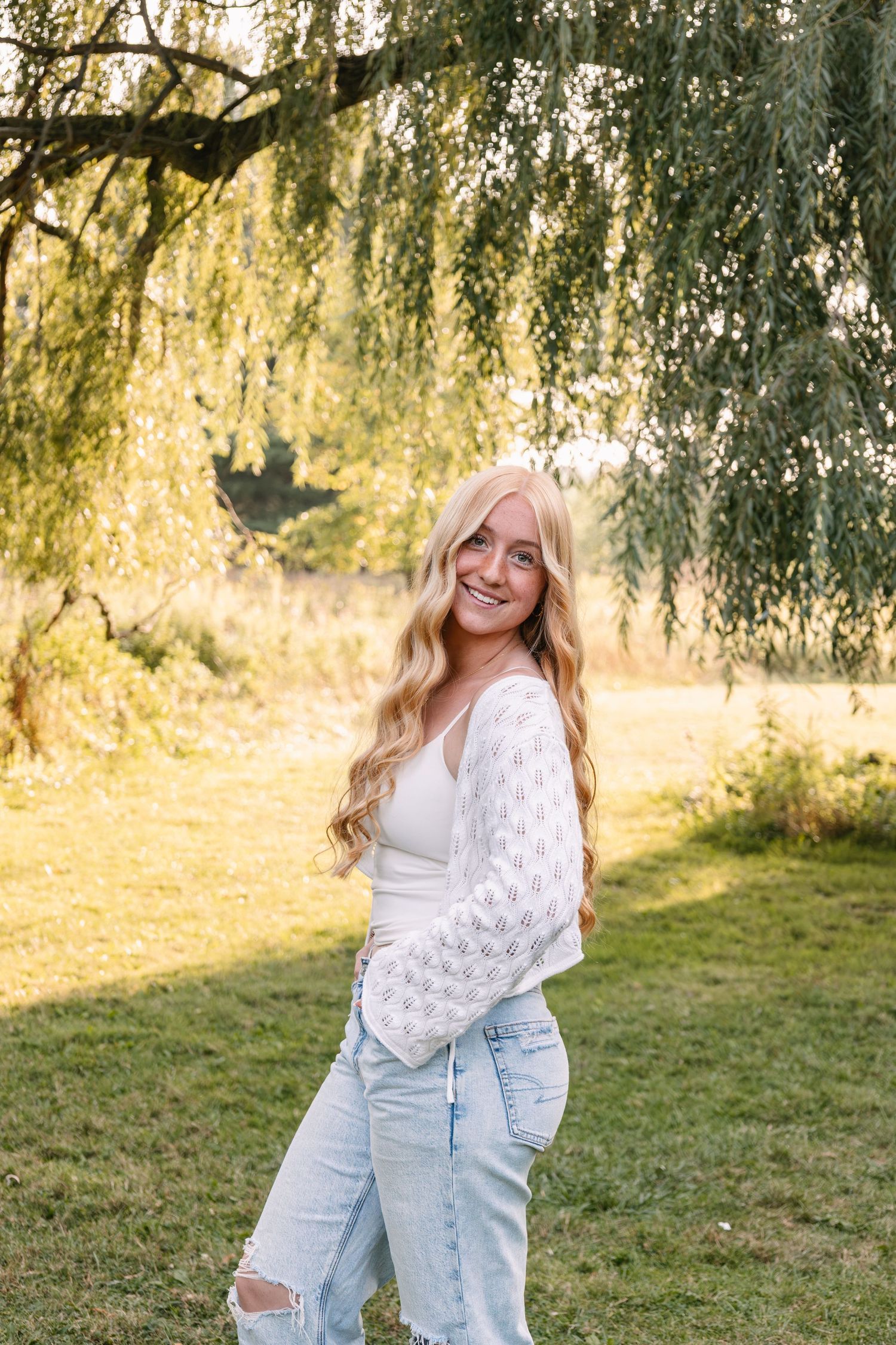 Young blonde woman smiling in white lace top and ripped jeans under a willow tree in a sunny green field.