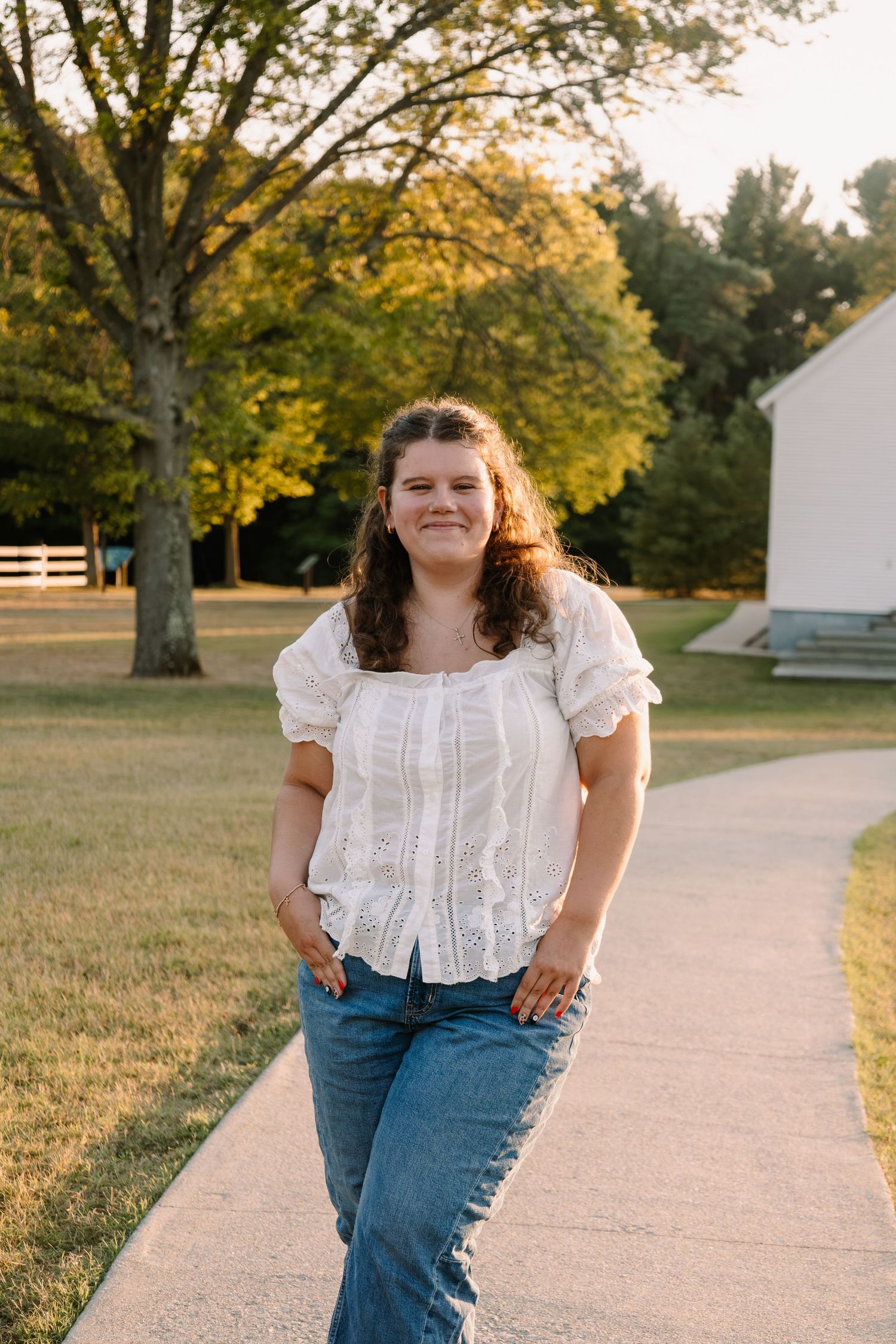 Young woman in white lace top and jeans smiling on a sunlit pathway surrounded by lush green trees.