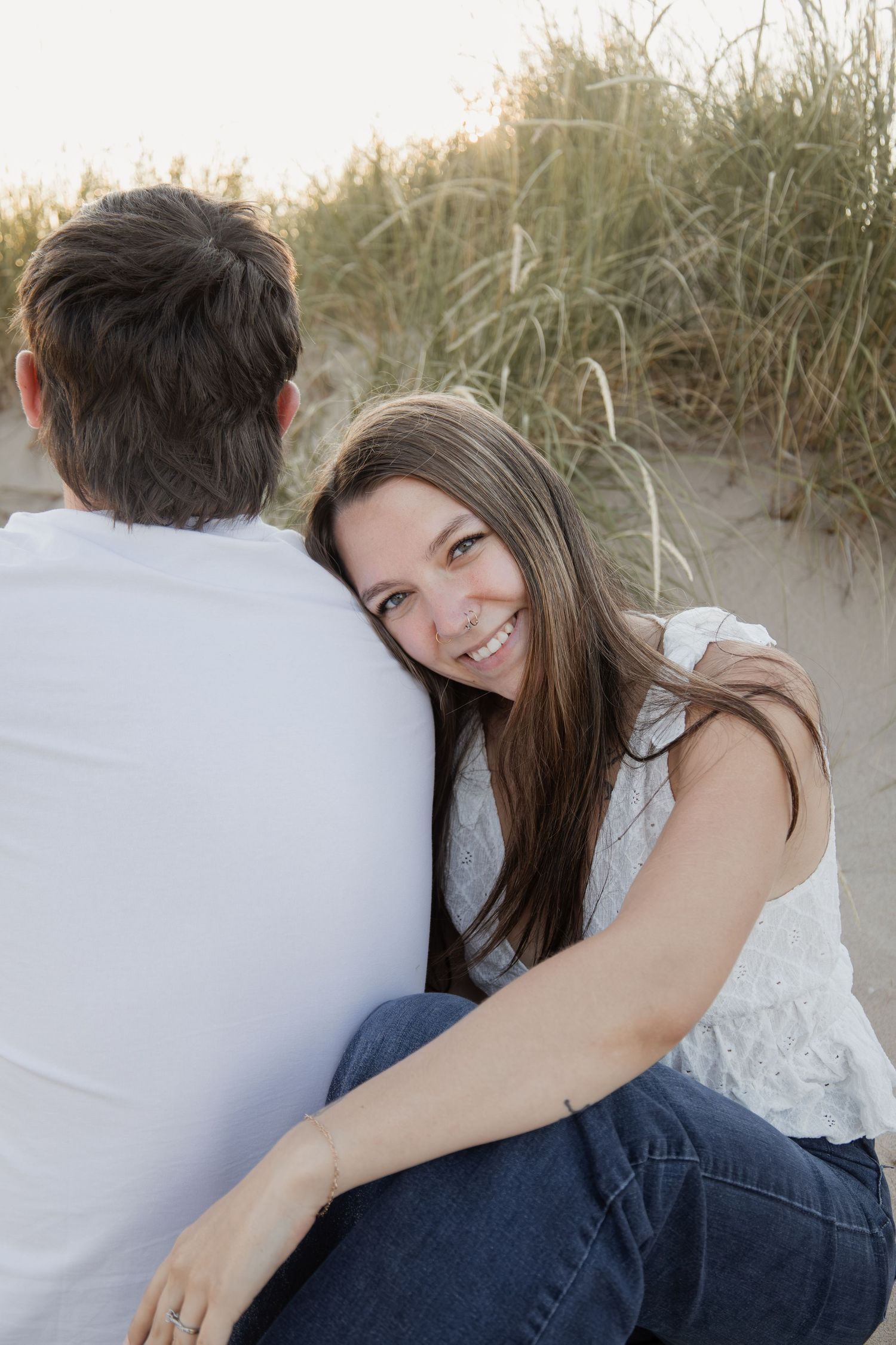 A smiling woman with long brown hair rests her head on a man's shoulder on a sandy beach.