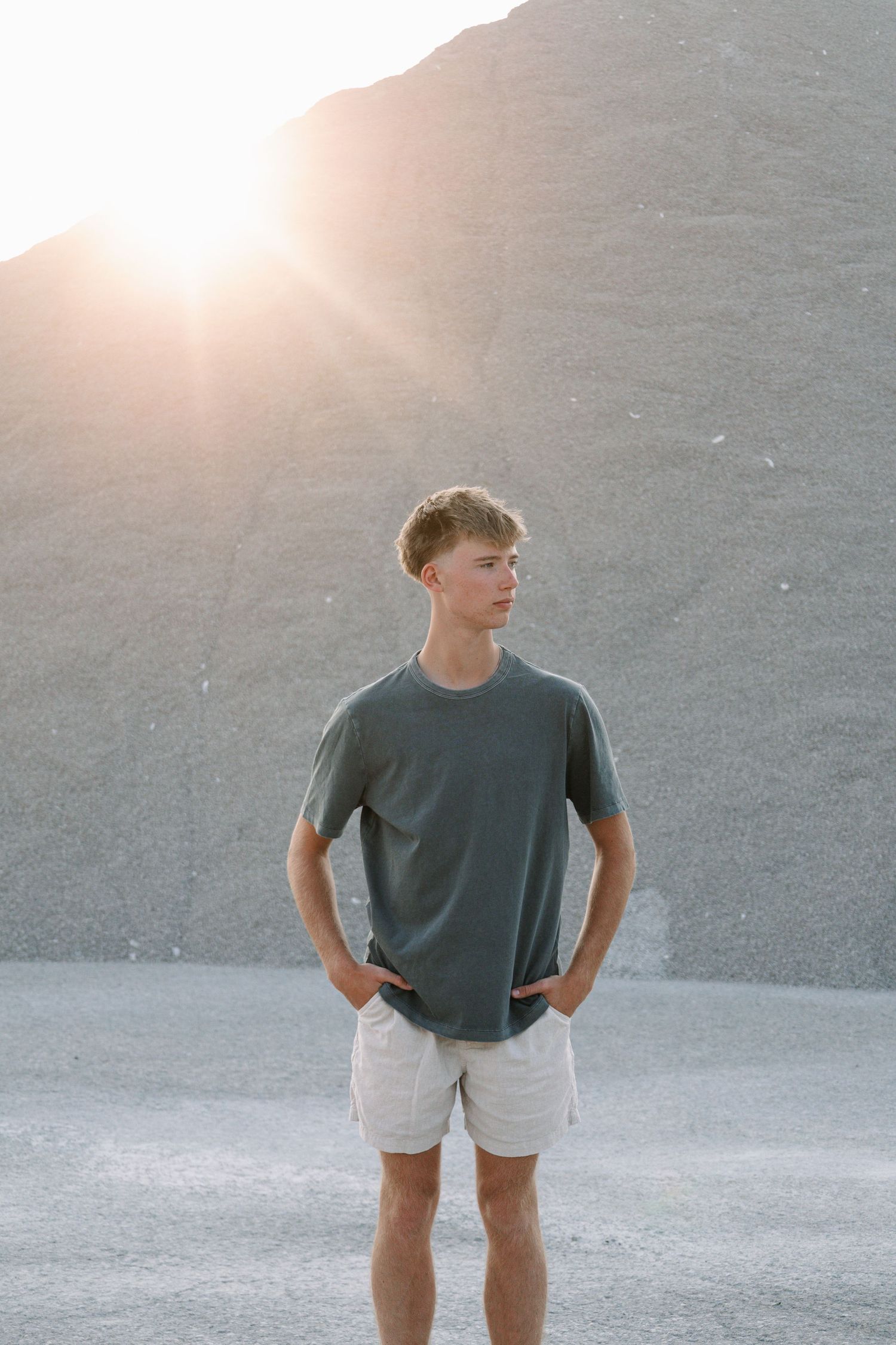 Young man in gray t-shirt and white shorts standing outdoors with sunlight glowing behind a rocky hillside.