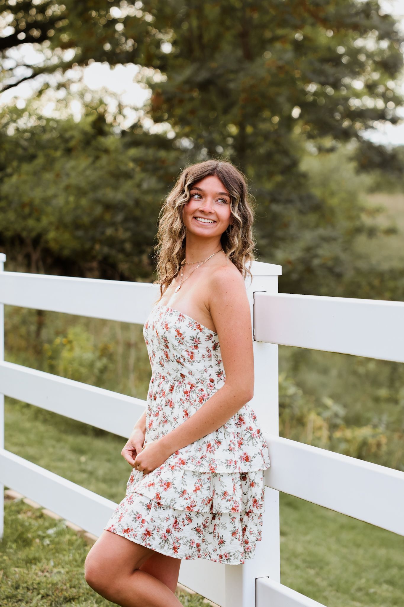 Young woman in floral dress leaning against white fence in garden.
