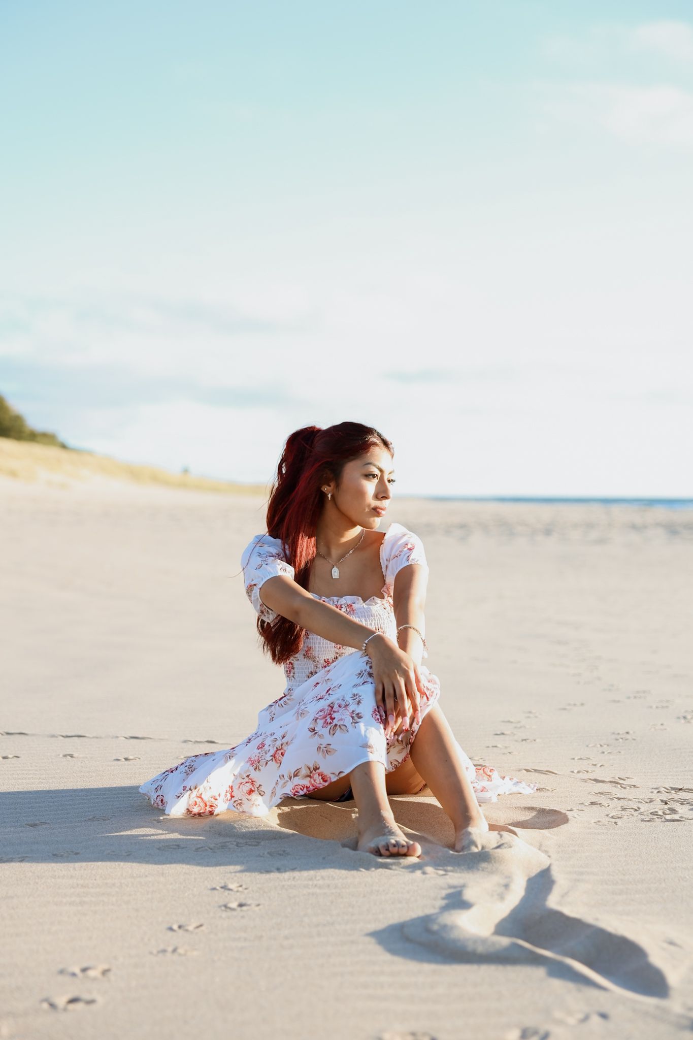 Young woman in floral dress sitting on wide sandy beach under blue sky.