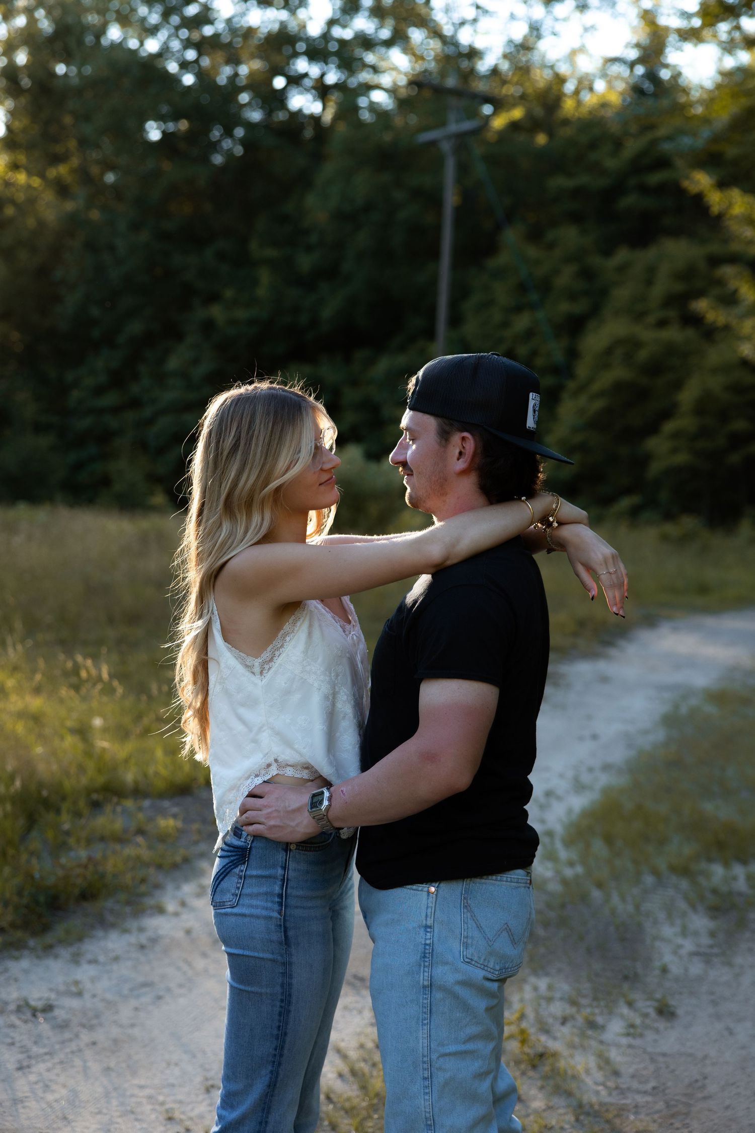 Couple embracing on a dirt path at golden hour, woman resting arms on mans shoulders, surrounded by trees.