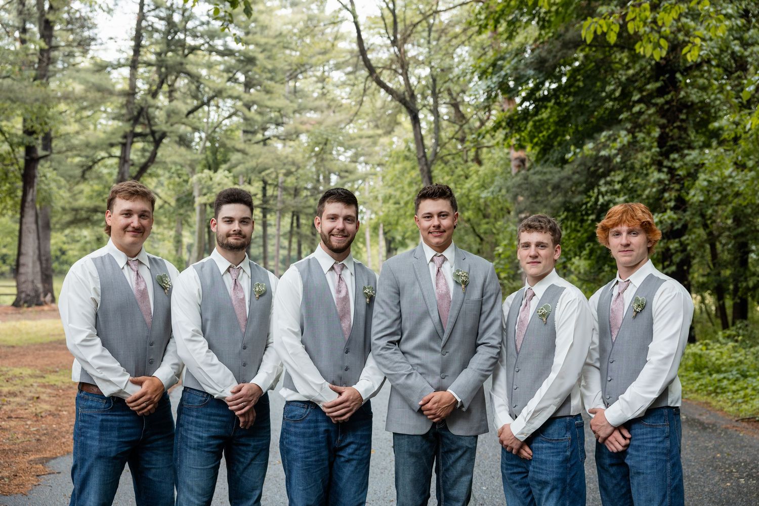 Groom and five groomsmen in gray vests, jeans, and pink ties pose outdoors among trees.