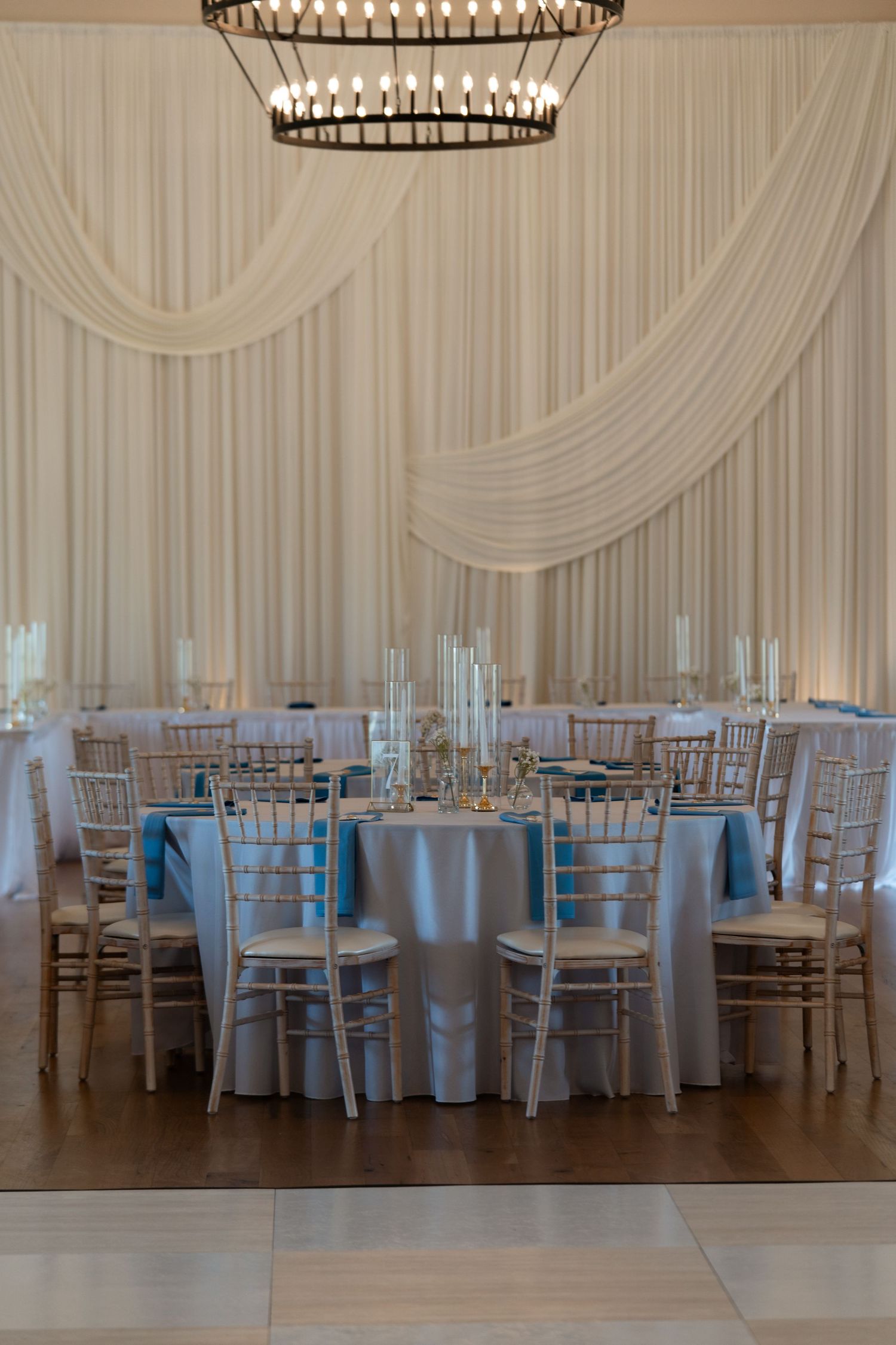 Elegant wedding reception head table with blue linens, white draping backdrop, and candlestick centerpieces.